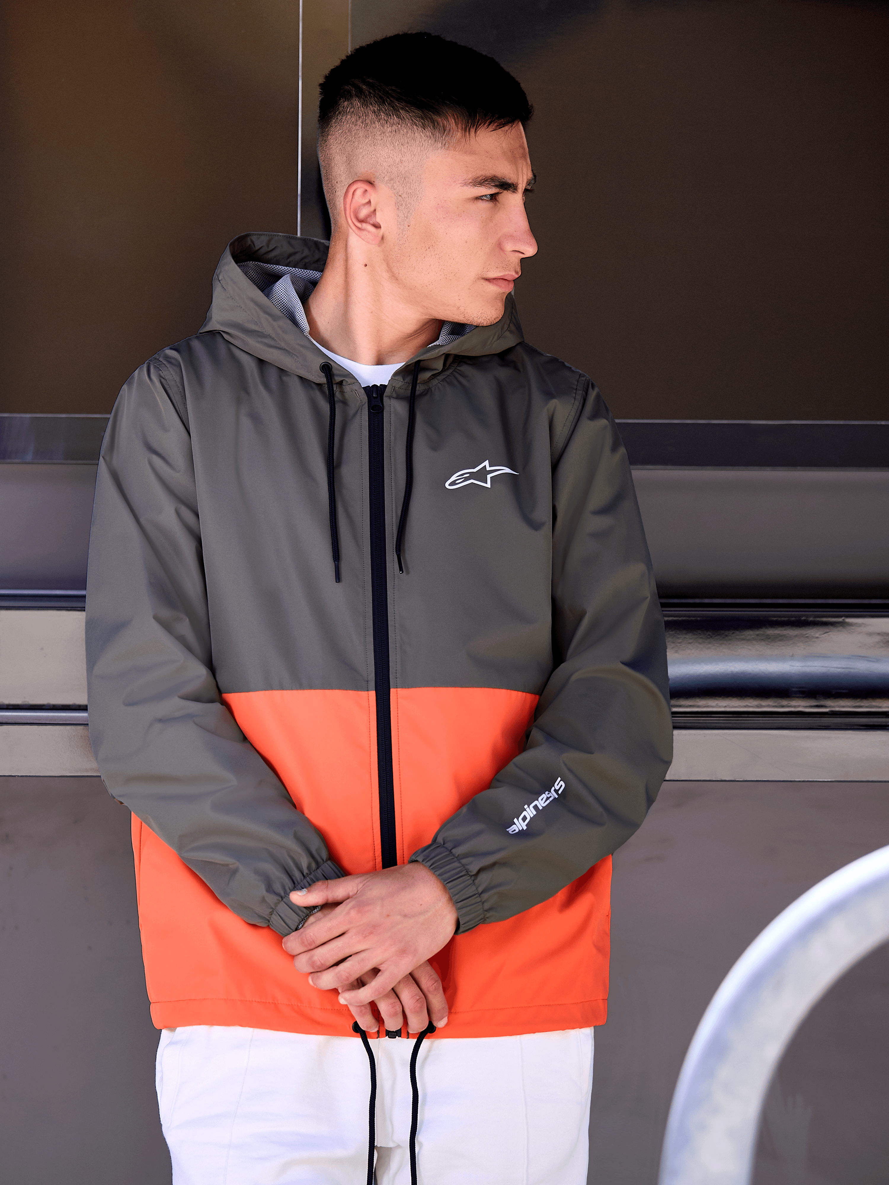 Man wearing the Alpinestars Speeder Windbreaker Jacket, Military Orange and Dark Green color-blocked design, hooded casual outerwear with white logos on chest and sleeve, paired with white pants for a motorsport-inspired lifestyle look