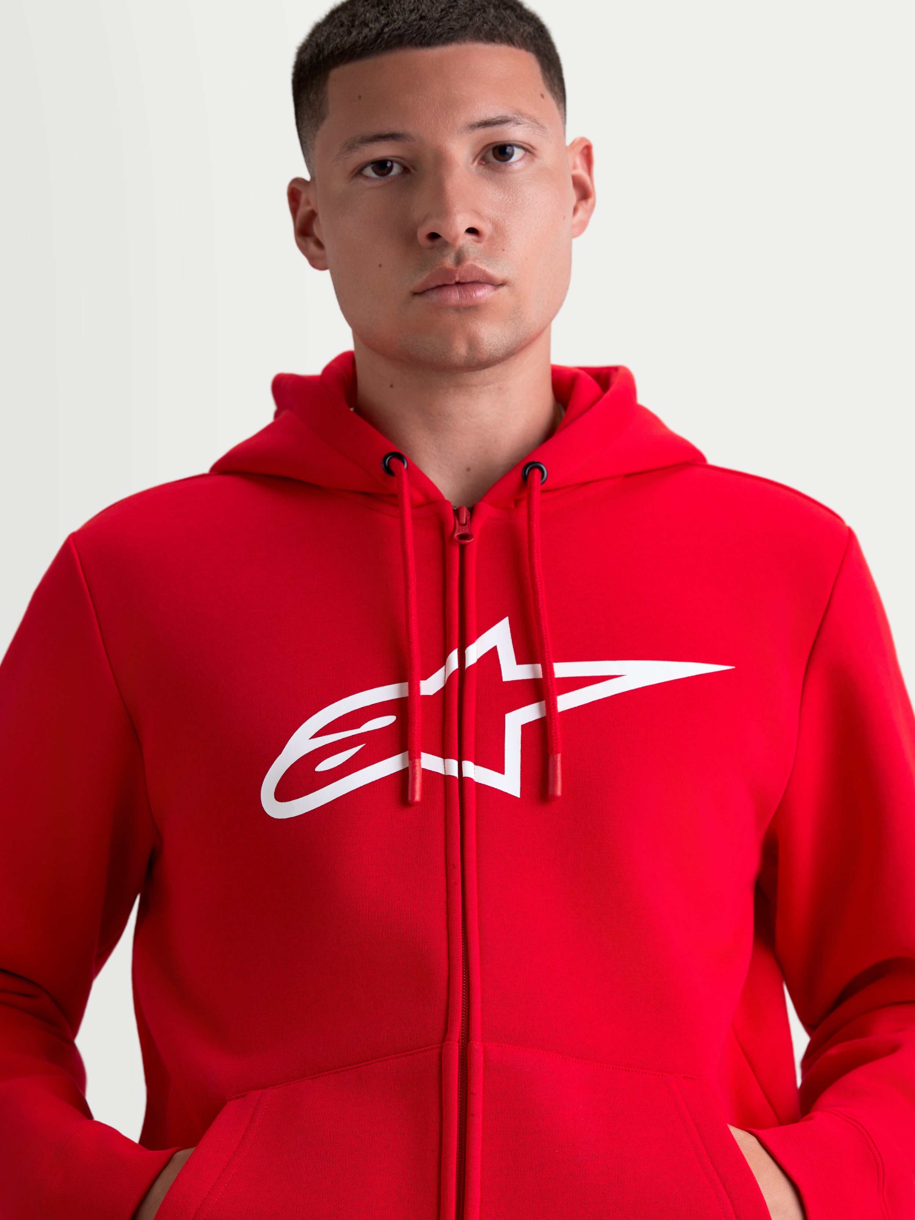 Alpinestars Ageless Zip V3 hættetrøje, rød sportswear-hættetrøje med gennemgående lynlås og hvidt A-star brystlogo, design med hætte og justerbare snore, vist på model for et motorsport-inspireret look.