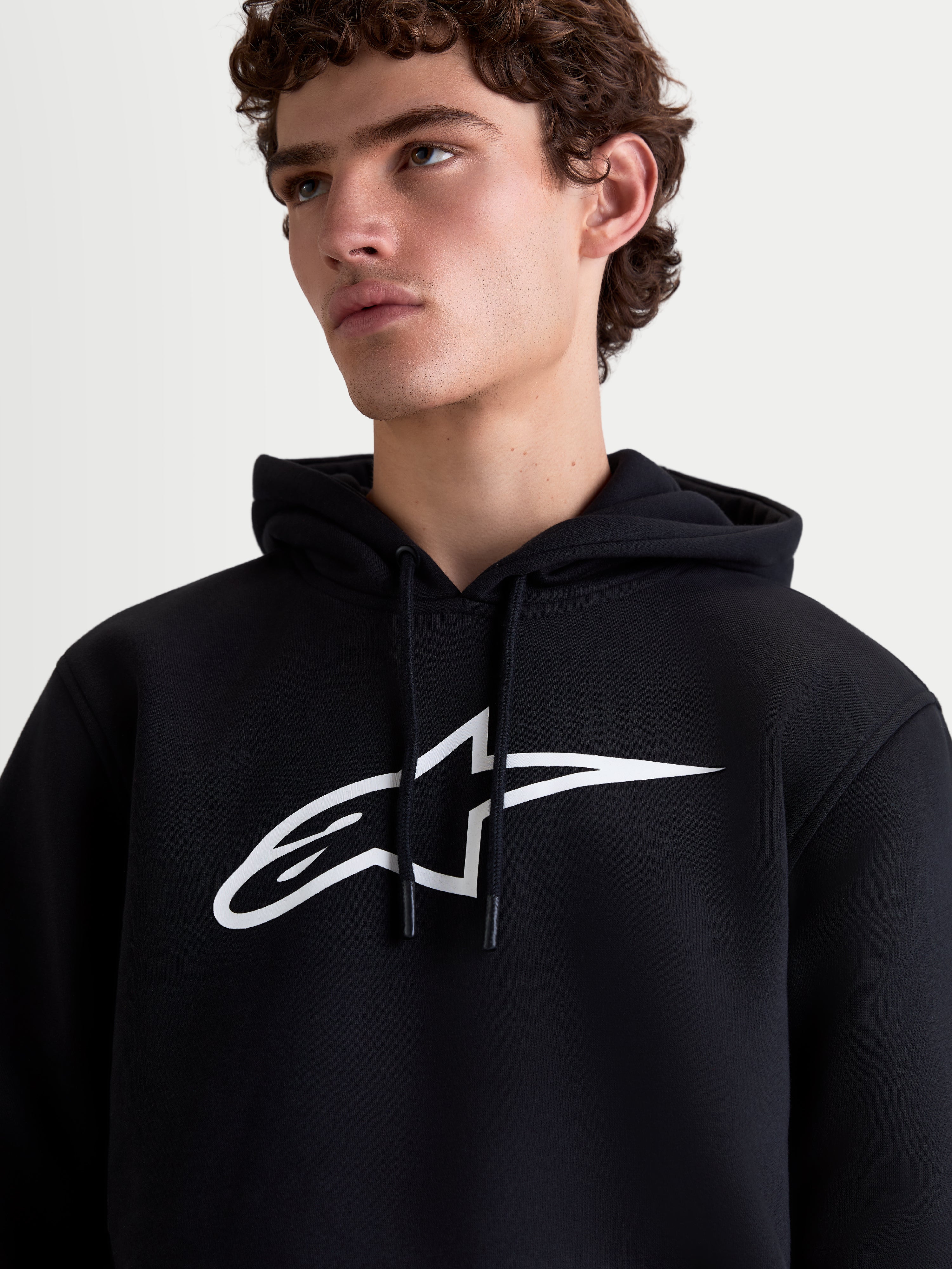 Alpinestars Ageless V3 hættetrøje, herre sportswear fleece, sort og hvid, tæt på-billede på model med et stort hvidt logo på brystet og hætte med snoretræk.