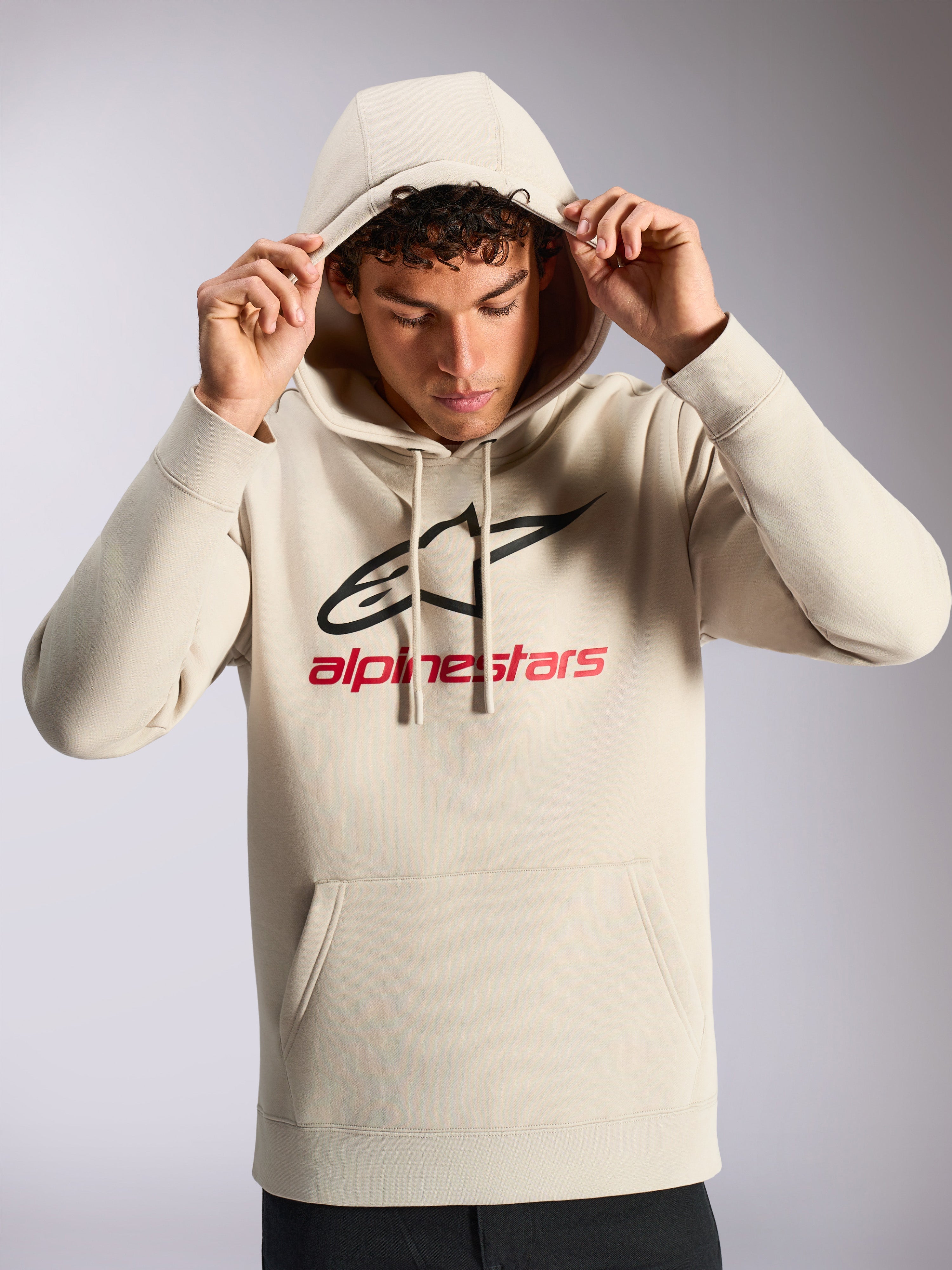 Alpinestars Always V3 hættetrøje, mandlig model iført off-white pullover fleece, sort og rødt lineært logo på fronten, hætte med snoretræk, kængurulomme, afslappet motorsport-inspireret streetwear