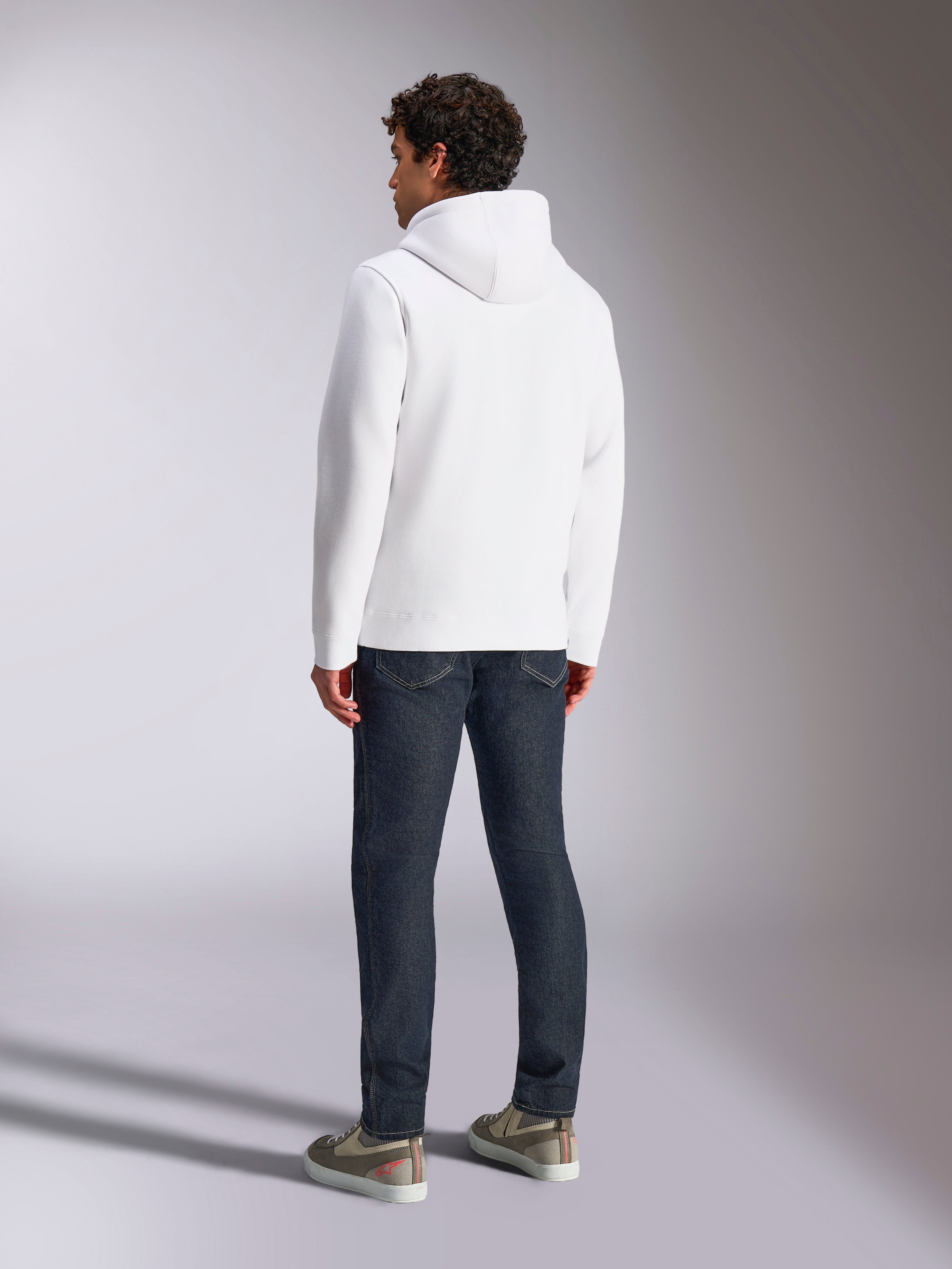 Alpinestars Blaze V3 Hættetrøje, mandlig model set bagfra iført hvid pullover-hættetrøje, afslappet sportstøj stylet med mørkeblå jeans og grå sneakers, moderne motorsport-inspireret design