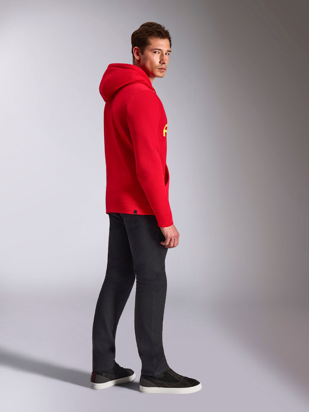 Alpinestars Ride V3 hættetrøje, afslappet sportstøj, rød med Hi-Vis gul branding, mandlig model iført rød hættetrøje med sorte bukser og sneakers, profilvisning fra siden, pullover fleece-design med ribkanter ved håndled og forneden.