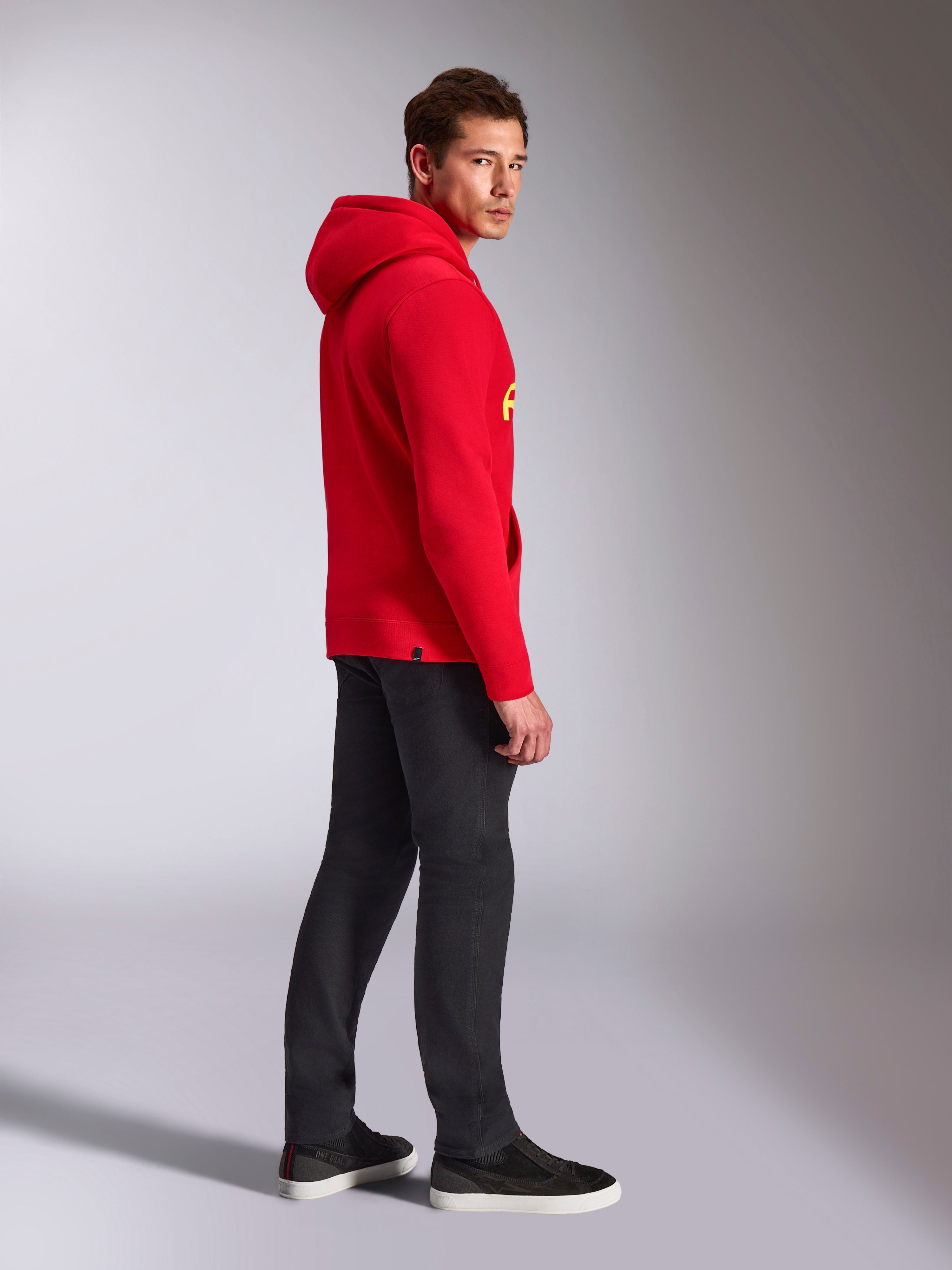 Alpinestars Ride V3 hættetrøje, afslappet sportstøj, rød med Hi-Vis gul branding, mandlig model iført rød hættetrøje med sorte bukser og sneakers, profilvisning fra siden, pullover fleece-design med ribkanter ved håndled og forneden.