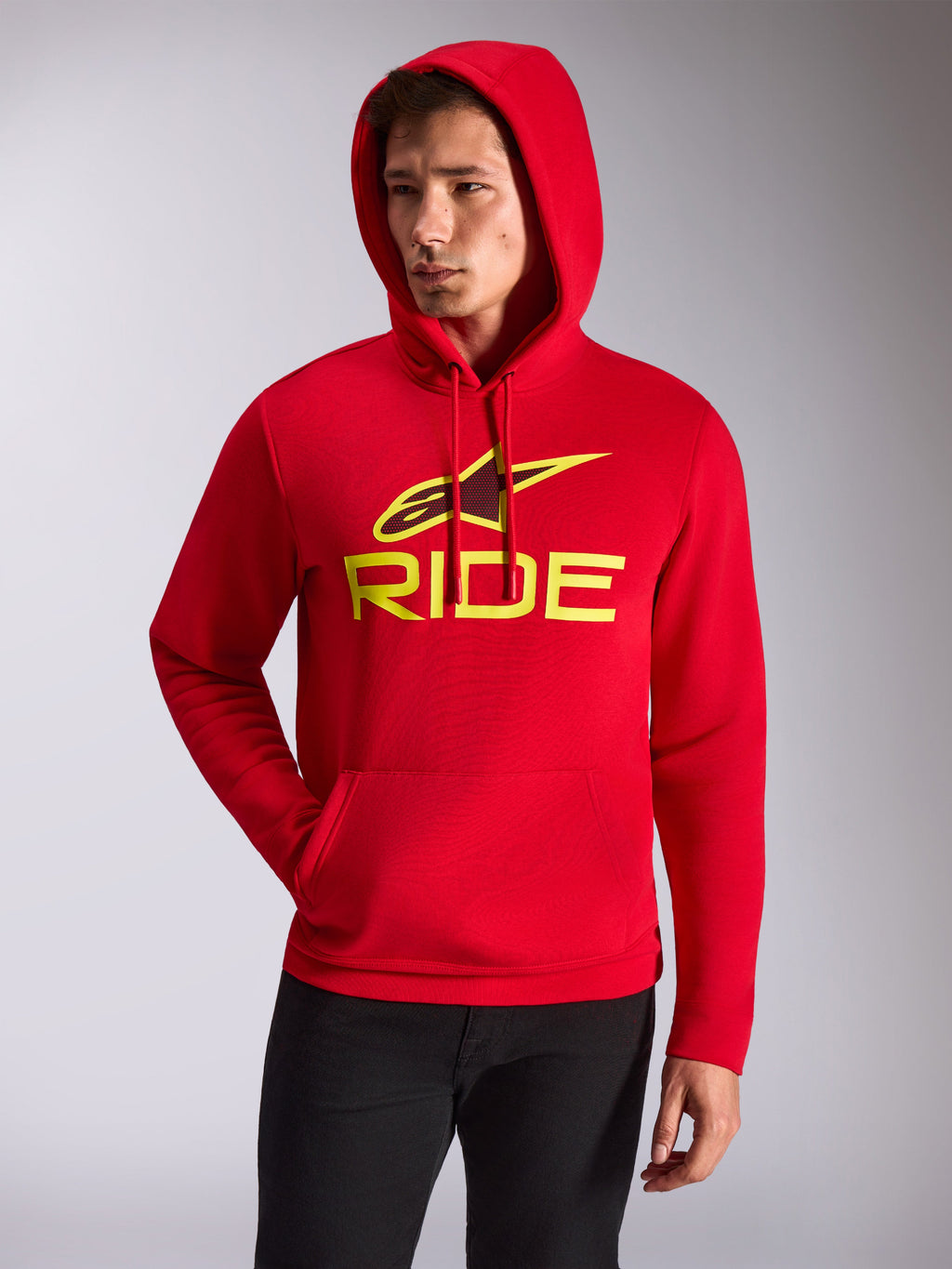 Alpinestars Ride V3 hættetrøje, mandlig model iført rød pullover-hættetrøje i fleece, hi-vis gult RIDE-logo og Alpinestars-stjerne på brystet, afslappet motorsport-streetwear, kængurulomme foran, båret med sorte bukser.