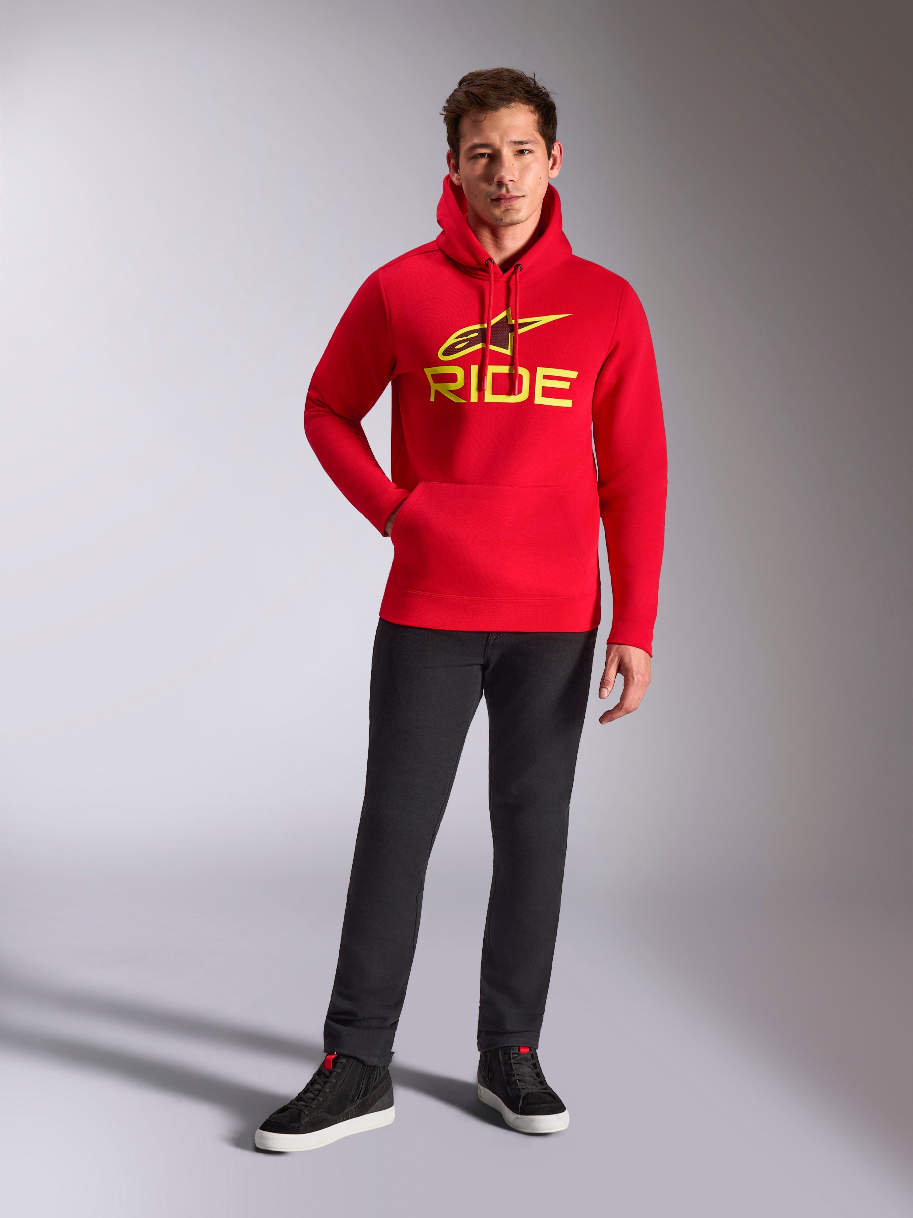 Alpinestars Ride V3 hættetrøje, rød pullover fleece, klart gult RIDE-logo med Astars-symbol på brystet, båret med sorte bukser og sorte high-top sneakers, casual motorsport-inspireret streetwear