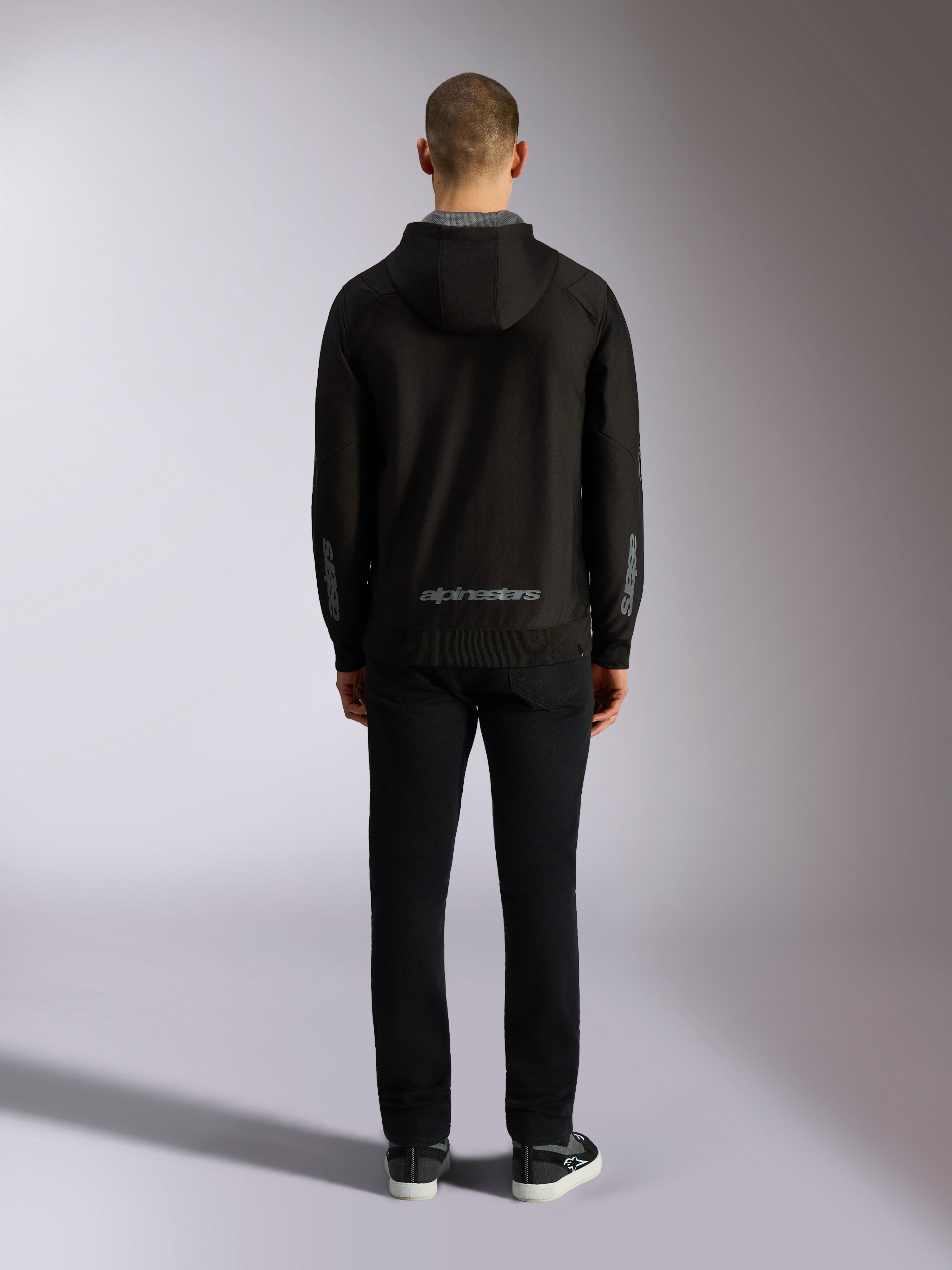 Set bagfra af Alpinestars Sessions V3 hættetrøje i Black Grey, med et gråt Alpinestars wordmark nederst på ryggen og astars-logoer på underarmene, båret af en model med sorte bukser, afslappet motorsport-inspireret streetwear