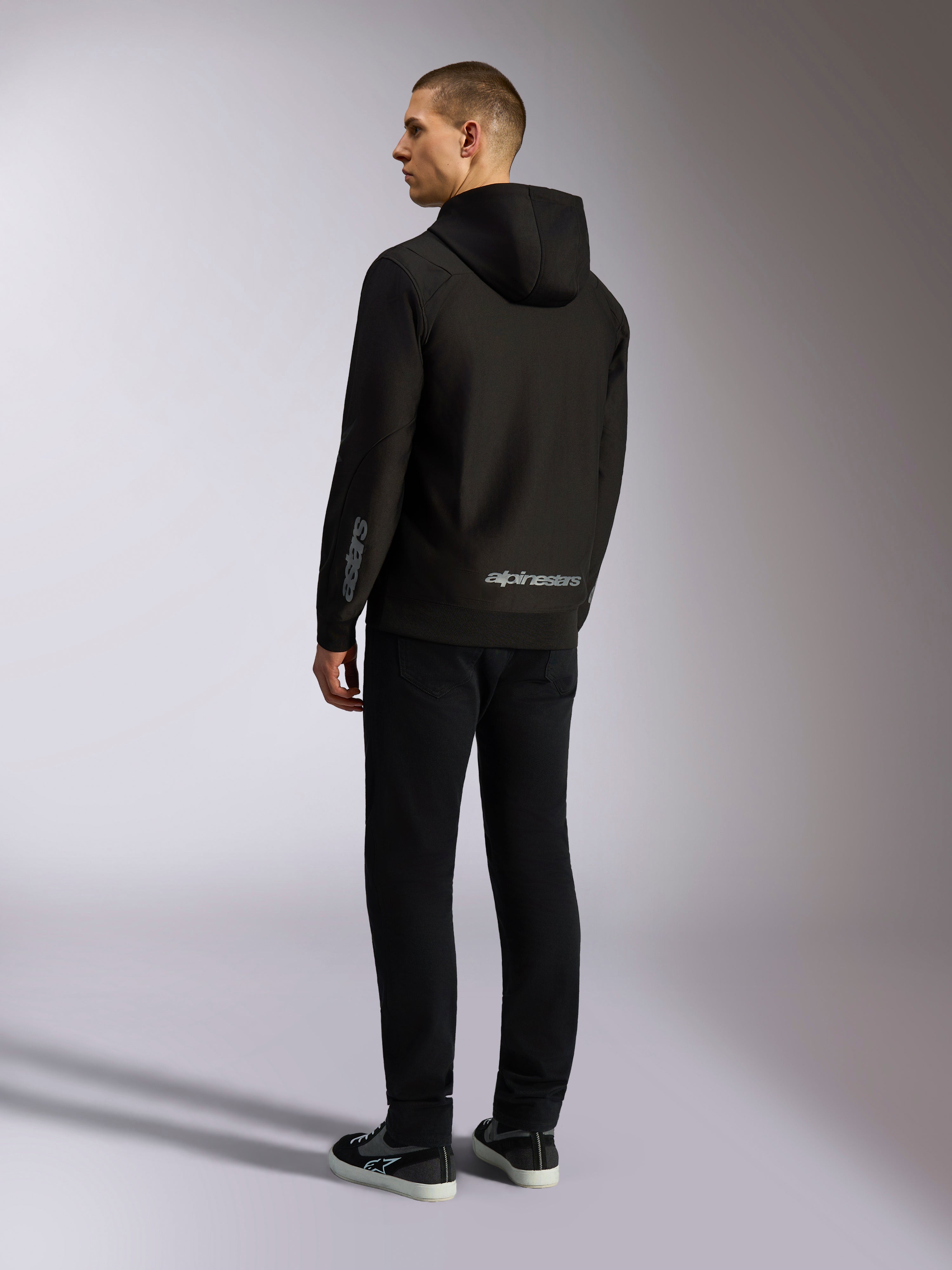 Alpinestars Sessions V3 hættetrøje, sort afslappet streetwear, set bagfra på en model, med gråt Alpinestars-navnetræk på den nederste del af ryggen og Astars-logo på ærmet, design med hætte og lynlås foran, moderne motorsport-inspireret styling