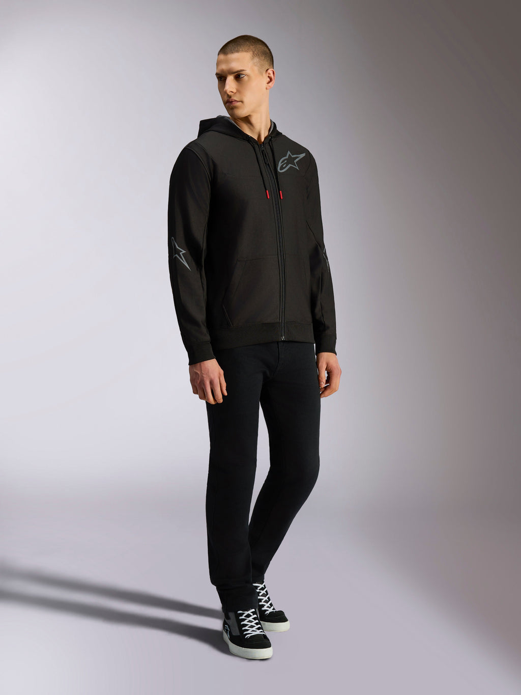 Alpinestars Sessions V3 hættetrøje, polyesterfleece med lynlås foran, sort og grå, grå logoer på bryst og ærmer, hætte med snoretræk med røde spidser, helkropsvisning af model i afslappet motorsport streetwear