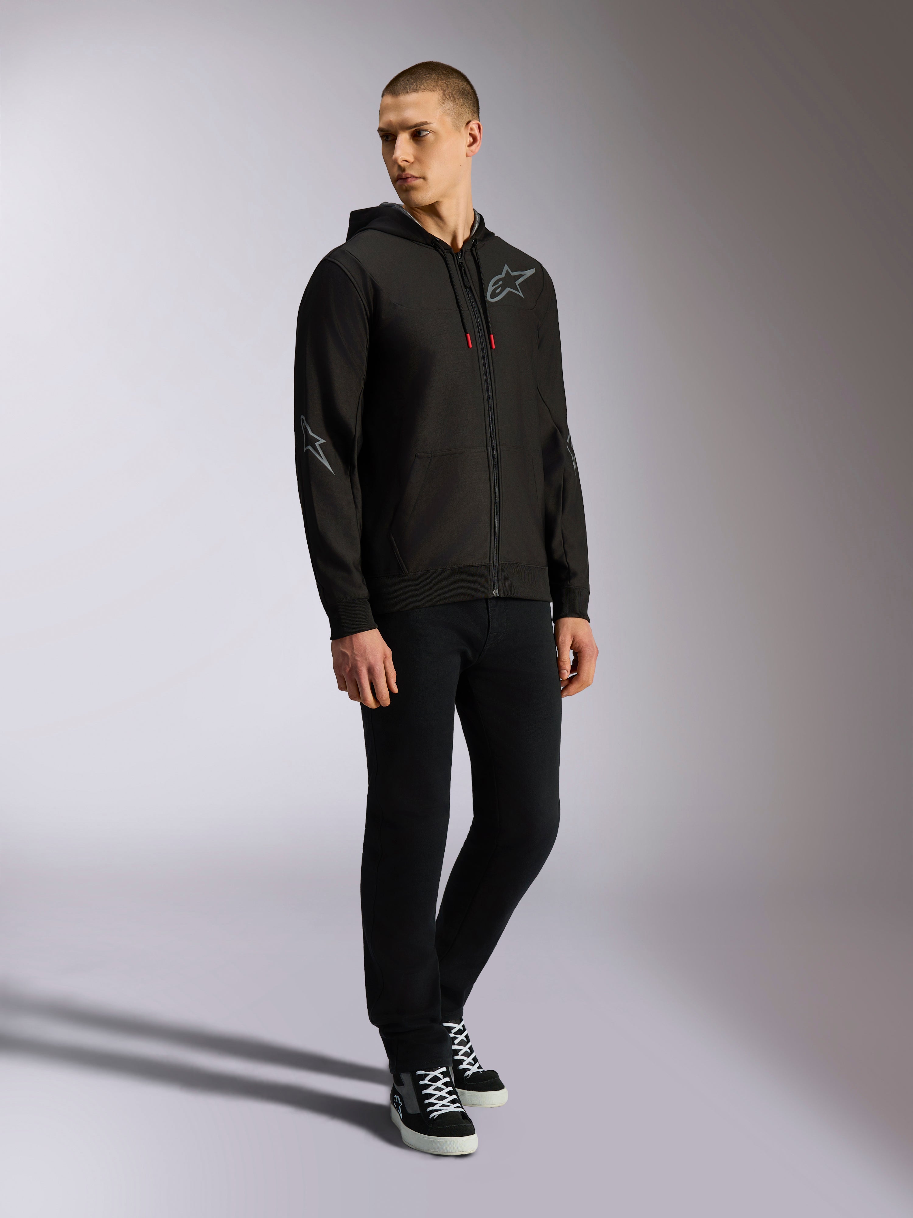 Alpinestars Sessions V3 hættetrøje, polyesterfleece med lynlås foran, sort og grå, grå logoer på bryst og ærmer, hætte med snoretræk med røde spidser, helkropsvisning af model i afslappet motorsport streetwear