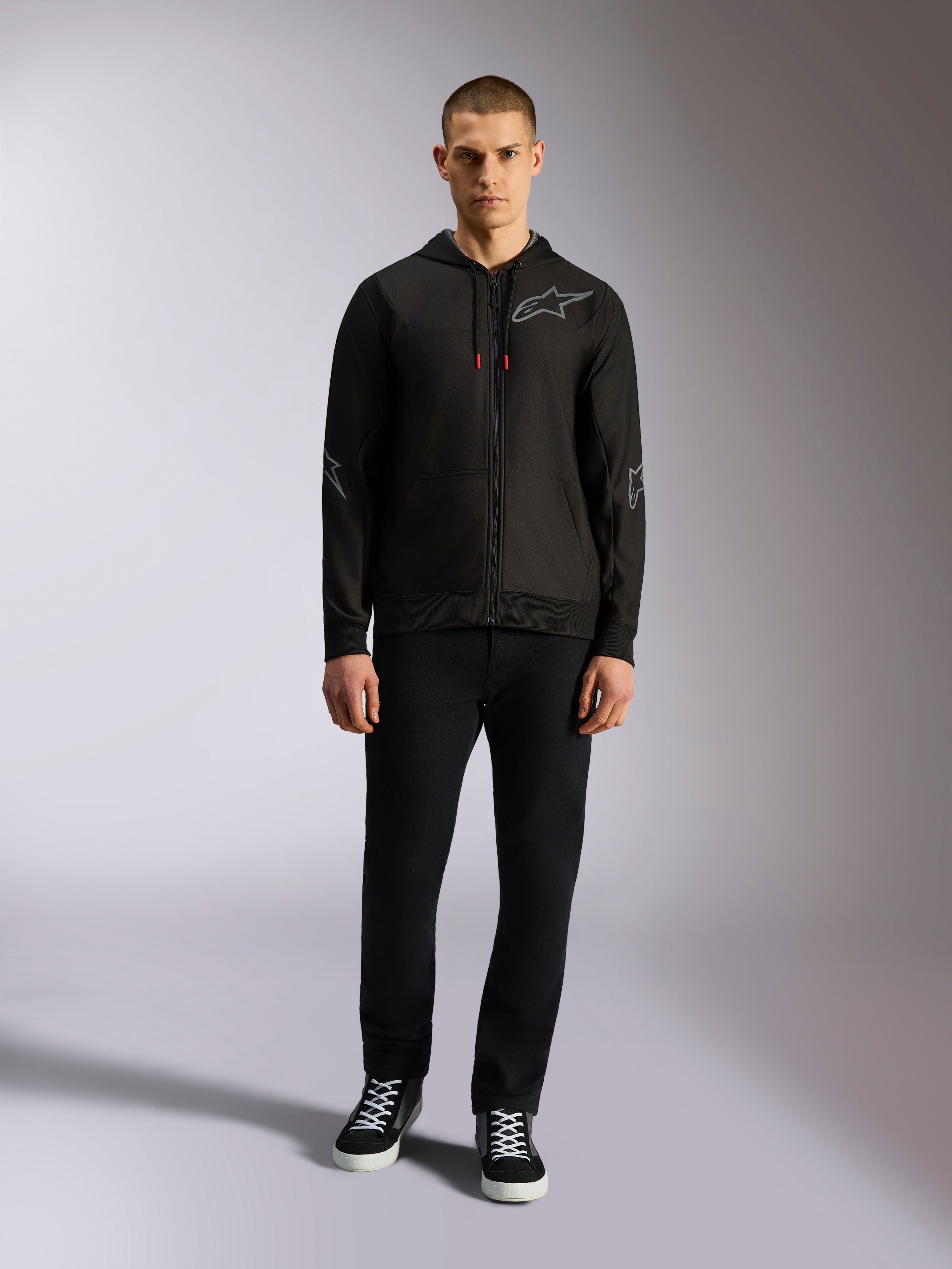 Alpinestars Sessions V3 hættetrøje, sort hættetrøje med lynlås, gråt Astars-logo på brystet, grafik på albuer og underarme, røde detaljer på snoretrækket, vist på mandlig model med sorte bukser og sneakers, afslappet motorsportstøj