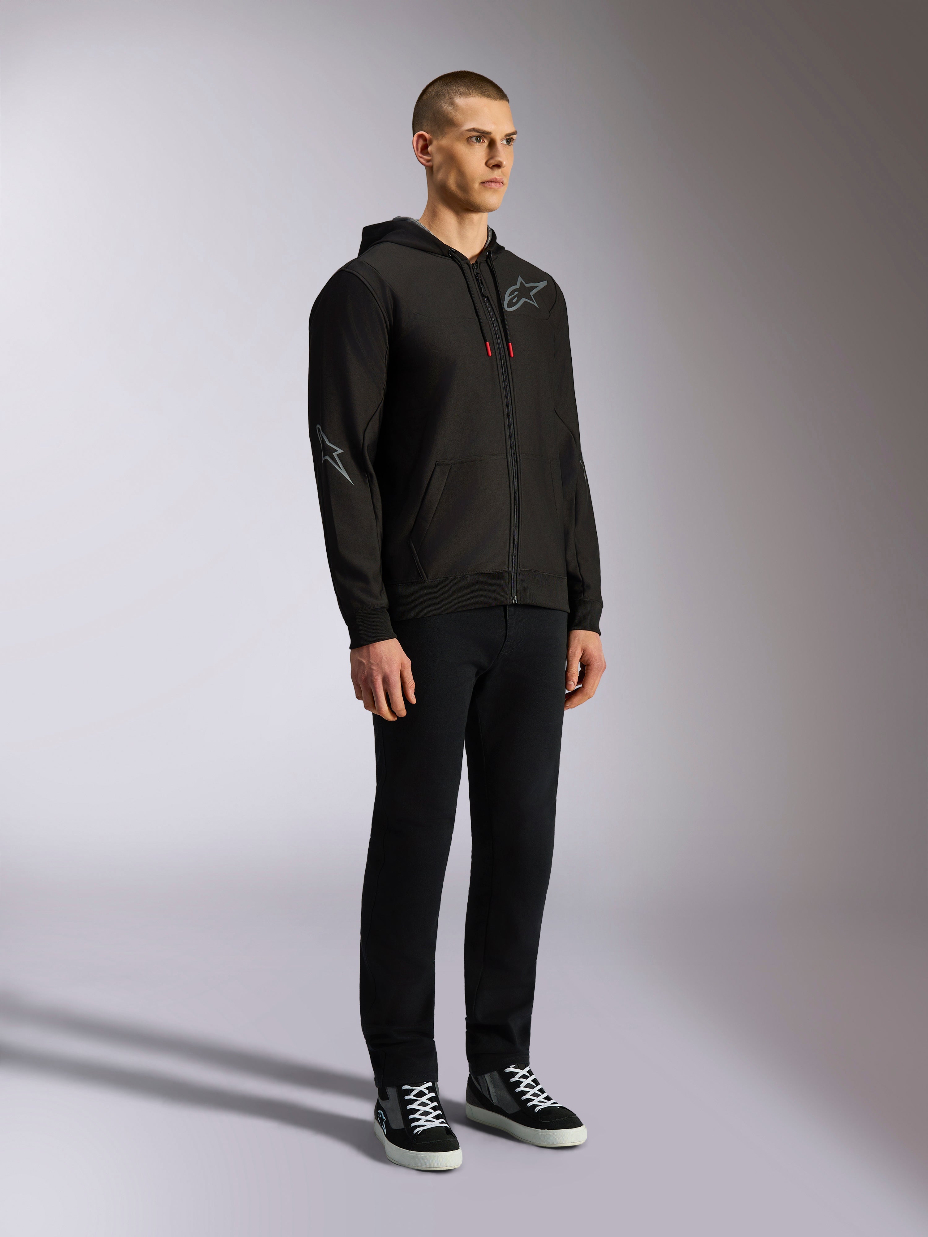 Alpinestars Sessions V3 hættetrøje, afslappet motorsport streetwear, sort hættetrøje med lynlås med gråt stjernelogo på brystet og ærmet, snoretræk med røde spidser, ribkanter på ærmer og bund, bæres af en model med sorte bukser og sneakers