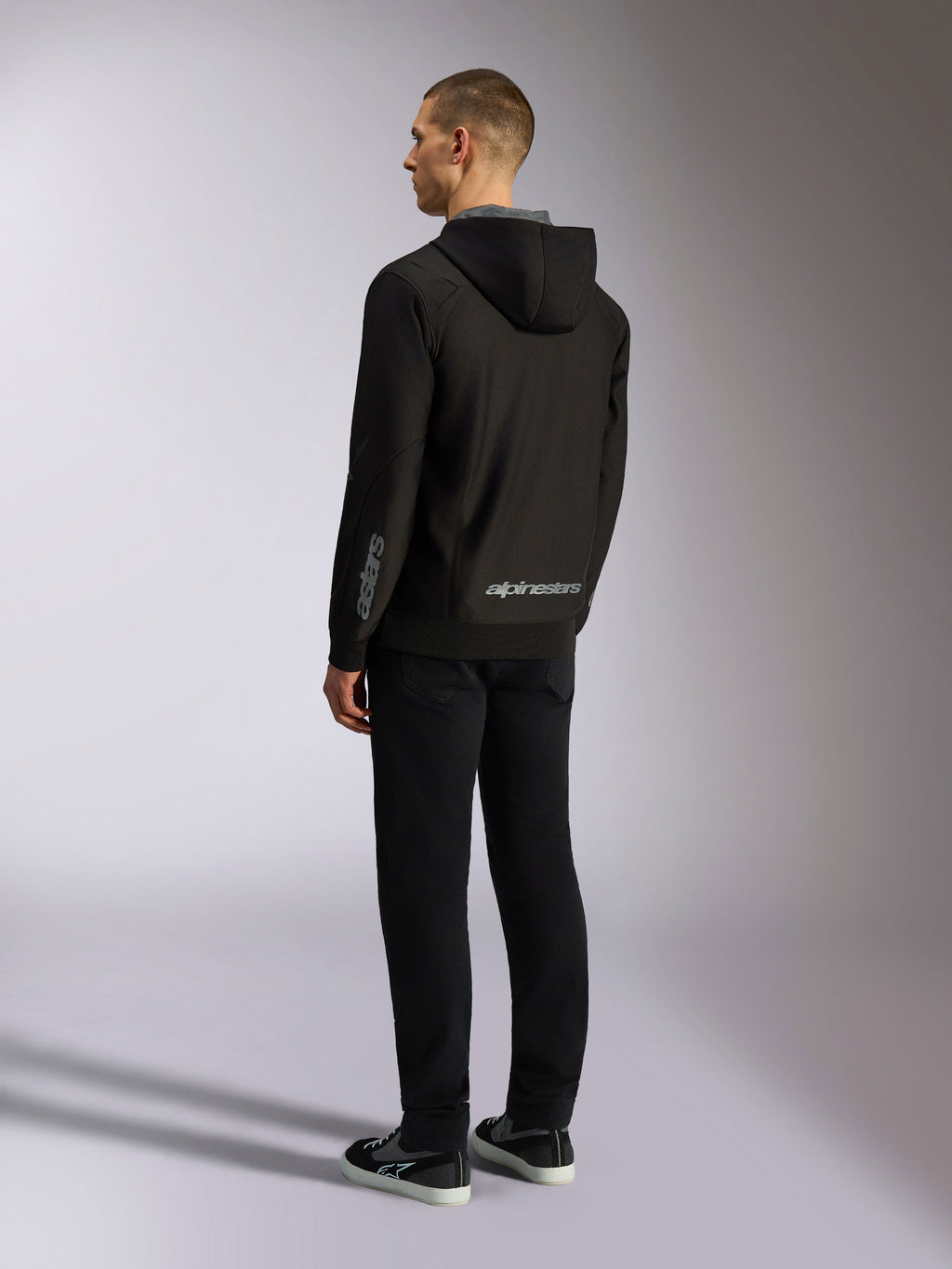 Alpinestars Sessions V3 hættetrøje, afslappet motorsport-streetwear, sort med gråt wordmark på den nederste del af ryggen og underarmen, polyfleece med lynlås foran og mesh-foret hætte, set bagfra på model, moderne atletisk design