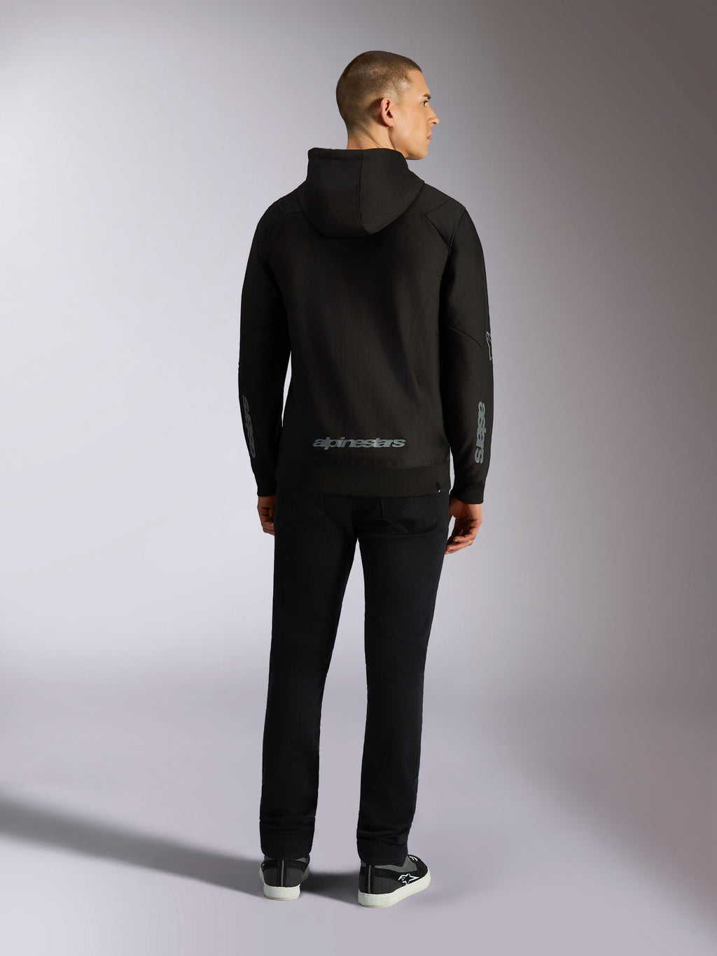 Alpinestars Sessions V3 hættetrøje, afslappet streetwear, sort med gråt Alpinestars-navnetræk på den nederste del af ryggen og 'astars'-branding på ærmerne, mandlig model set bagfra iført hættetrøje og sorte bukser, moderne motorsport-inspireret design