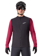 Alpinestars A-Dura Thermal Vest, sort isoleret cykelvest, båret af en model med en bordeauxrød langærmet trøje, sort MTB-hjelm og solbriller, hvidt logo på brystet, letvægts termisk beskyttelse til mountainbiking