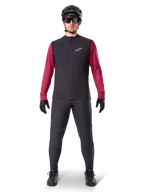 Alpinestars A-Dura Thermal Vest, sort cykelvest båret over en bordeauxfarvet langærmet trøje, kombineret med mørkegrå MTB-bukser, sort hjelm og solbriller, teknisk mountainbike-beklædning til kørsel i koldt vejr.