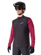 Alpinestars A-Dura Thermal Vest, sort termisk cykelvest, bæres over en vinrød langærmet trøje, med hvidt logo på brystet, kombineret med en sort MTB-hjelm og solbriller, isolerende præstationslag til mountainbiking