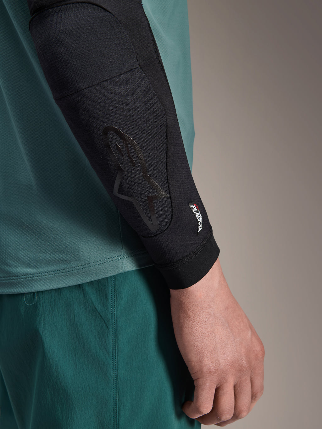 Alpinestars A-Motion Plasma Elbow Sleeve, sort MTB-albuebeskytter, nærbillede båret på armen med blågrøn jersey, med tonalt Astars-logo og Nucleon Plasma-branding, slankt mesh-design til off-road cykling