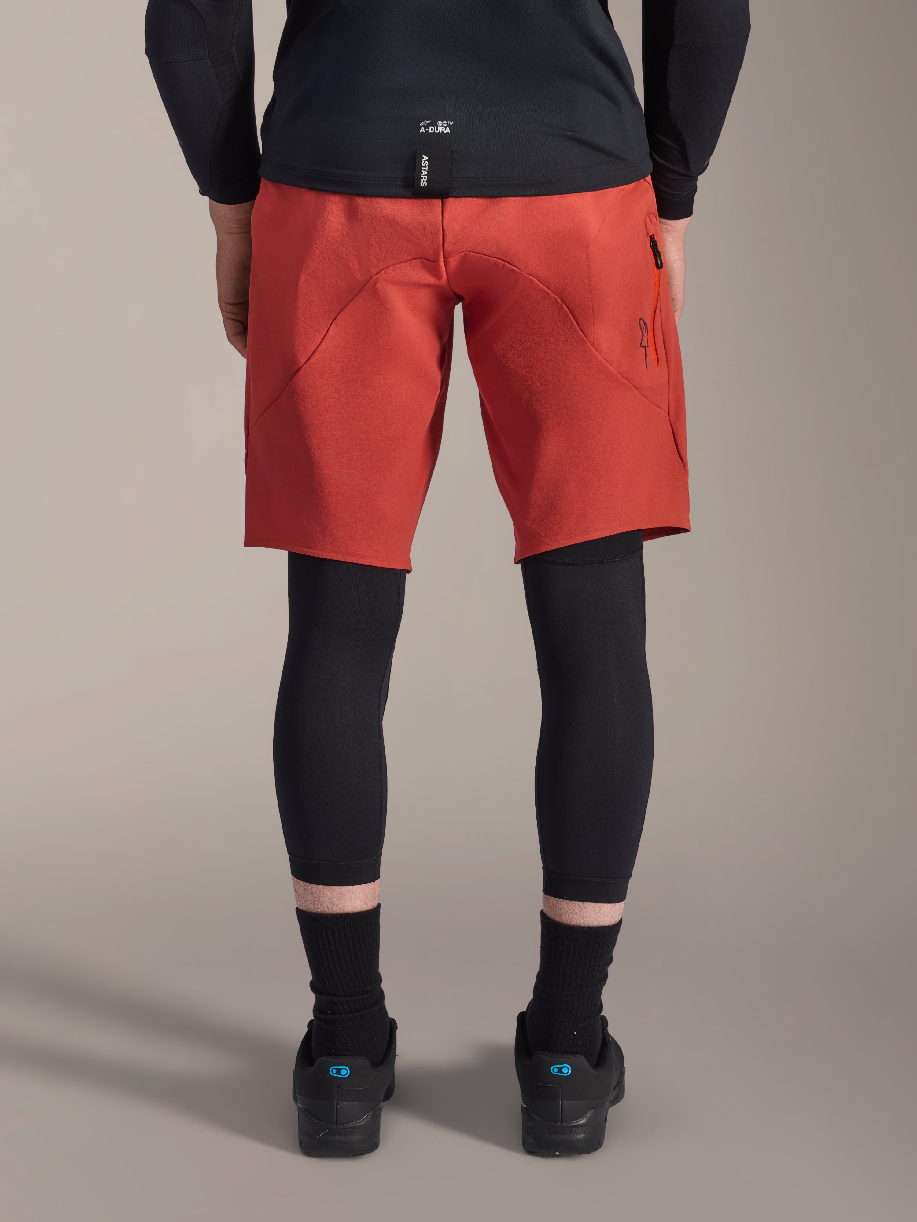 Alpinestars A-Motion Plasma knæærme, sorte MTB-knæbeskyttere, set bagfra båret under røde mountainbike-shorts med en sort jersey og tekniske cykelsko, beskyttelsesudstyr til mountainbike