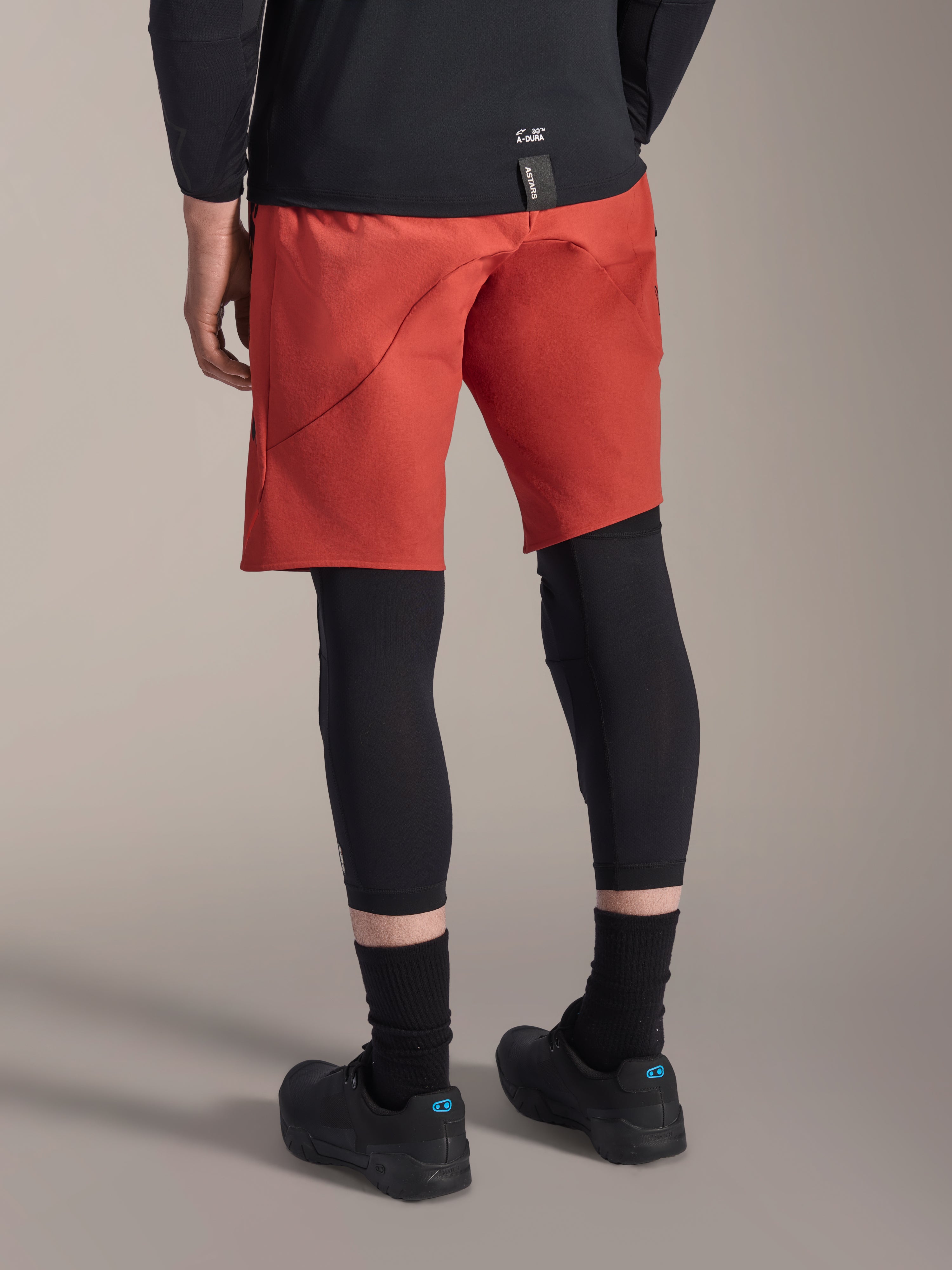Alpinestars A-Motion Plasma Knæærme, sort MTB knæbeskytter, båret med røde mountainbike-shorts og sorte cykelsko, set bagfra viser det lavprofilerede slip-on design til terrænkørsel
