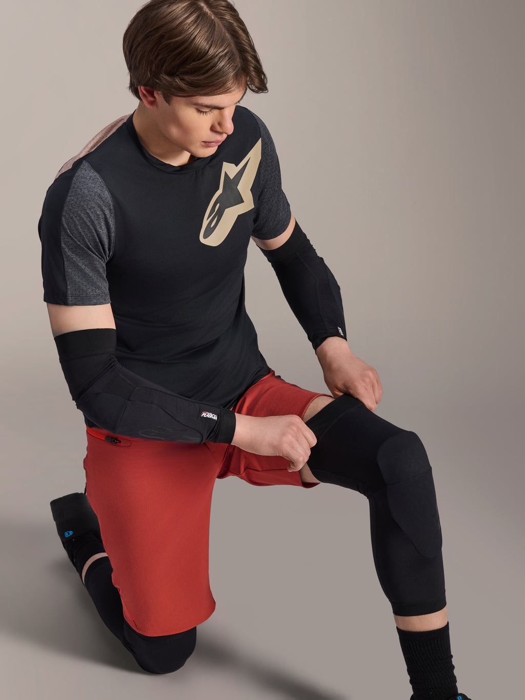 Alpinestars A-Motion Plasma knæærme, MTB knæbeskytter, sort, båret af en rytter i knælende stilling, kombineret med en sort og grå t-shirt med guldlogo, røde shorts og sorte albuebeskyttere, professionelt mountainbike-beskyttelsesudstyr