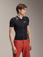 Mandlig model iført Alpinestars A-Motion Plasma beskyttelsesjakke, kortærmet, Black Camo med grå camouflage-skulderpaneler, med integrerede skulder- og brystbeskyttere, hvidt stjernelogo på brystet, åndbart mesh-stof, kombineret med røde bukser