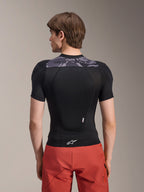 Alpinestars A-Motion Plasma beskyttelsesjakke - kortærmet, sort med grå camo-detaljer på den øverste del af ryggen, set bagfra med integreret rygskjold og hvidt Astars-logo ved kanten, kombineret med røde shorts, teknisk mountainbike-beskyttelsesudstyr