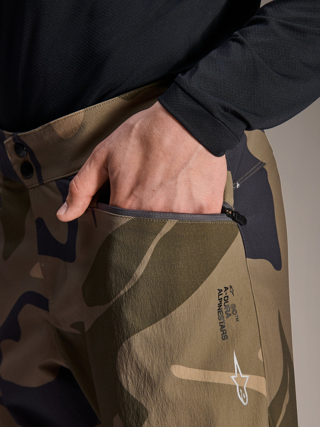Nærbillede af Alpinestars A-Dura Camo bukser, brune camouflage mountainbikebukser, med en hånd i en lynlåslomme, dobbelt trykknapslukning i taljen og Alpinestars-branding på låret.