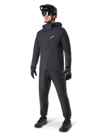 Mandlig mountainbiker iført Alpinestars A-Dura Waterproof Pants i sort, kombineret med en matchende sort vandtæt jakke med hætte, sort MTB-hjelm med visir, mørke cykelbriller og sorte handsker, komplet professionelt udstyrssæt til trail-kørsel i al slags vejr