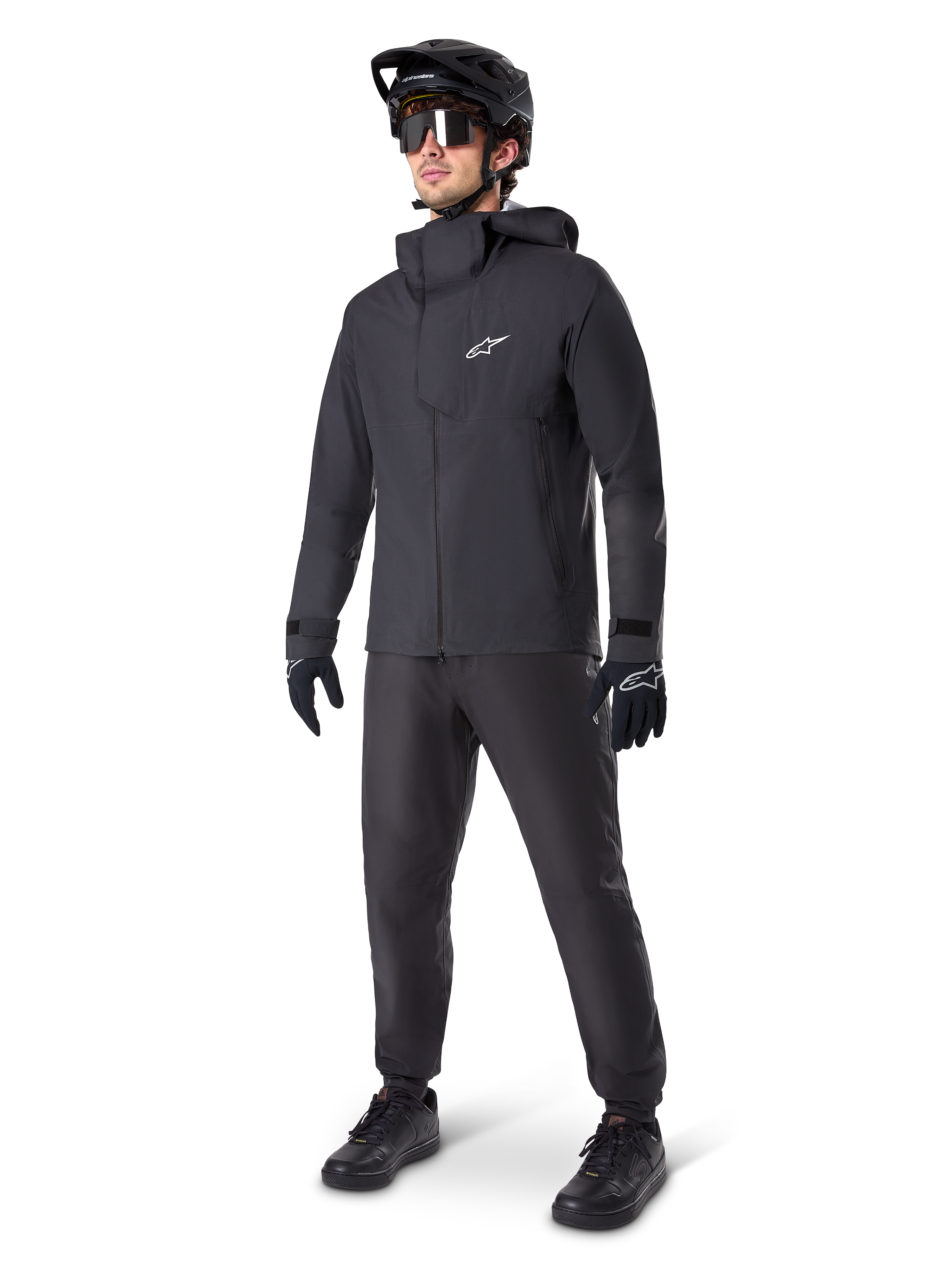 Mandlig mountainbiker iført Alpinestars A-Dura Waterproof Pants i sort, kombineret med en matchende sort vandtæt jakke med hætte, sort MTB-hjelm med visir, mørke cykelbriller og sorte handsker, komplet professionelt udstyrssæt til trail-kørsel i al slags vejr