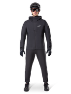 Alpinestars A-Dura Waterproof Pants, sorte MTB-bukser, båret som en del af et komplet helt sort mountainbike-udstyrssæt inklusiv en teknisk vandtæt jakke, sort hjelm, solbriller og handsker, designet til terrænkørsel i vådt vejr.