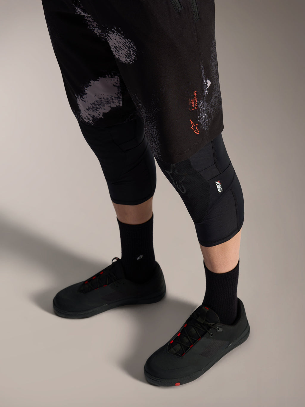 Alpinestars A-Dura Ride shorts i Black Camo med orange logo, vist med sorte knæbeskyttere og cykelsko til MTB