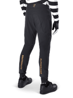 Alpinestars A-Aria Elite bukser, sorte MTB-bukser, set bagfra, med lodret guld Alpinestars-branding bag på benene, båret med sort- og hvidstribet trøje og sorte sko, performance-cykeltøj til mountainbike