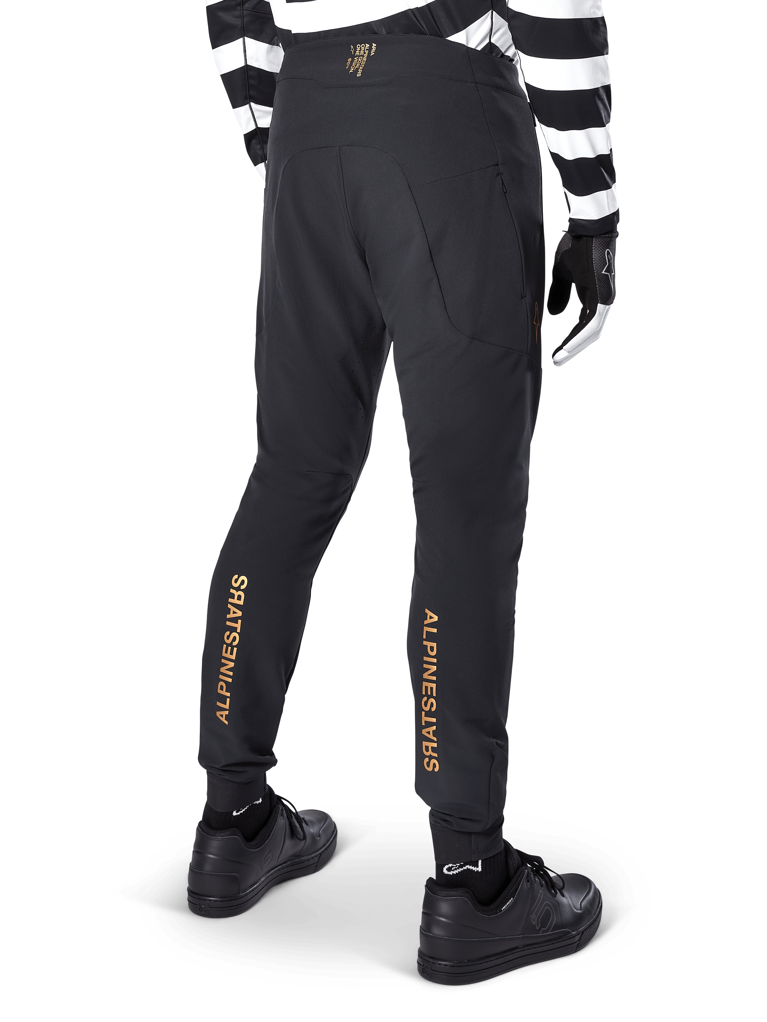 Alpinestars A-Aria Elite bukser, sorte MTB-bukser, set bagfra, med lodret guld Alpinestars-branding bag på benene, båret med sort- og hvidstribet trøje og sorte sko, performance-cykeltøj til mountainbike
