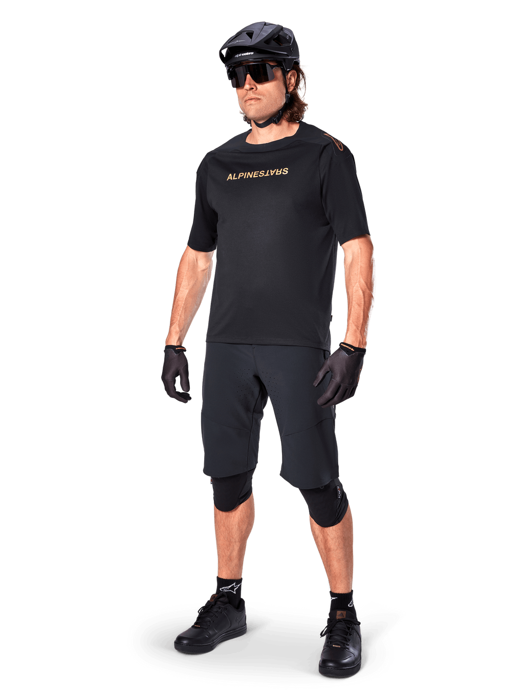 Cyklist iført Alpinestars A-Aria Elite shorts, sorte mountainbike-shorts, kombineret med en matchende sort trøje med guld-branding, sort hjelm, solbriller og knæbeskyttere, komplet performance-outfit til terrænkørsel