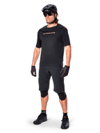Cyklist iført Alpinestars A-Aria Elite shorts, sorte mountainbike-shorts, kombineret med en matchende sort trøje med guld-branding, sort hjelm, solbriller og knæbeskyttere, komplet performance-outfit til terrænkørsel