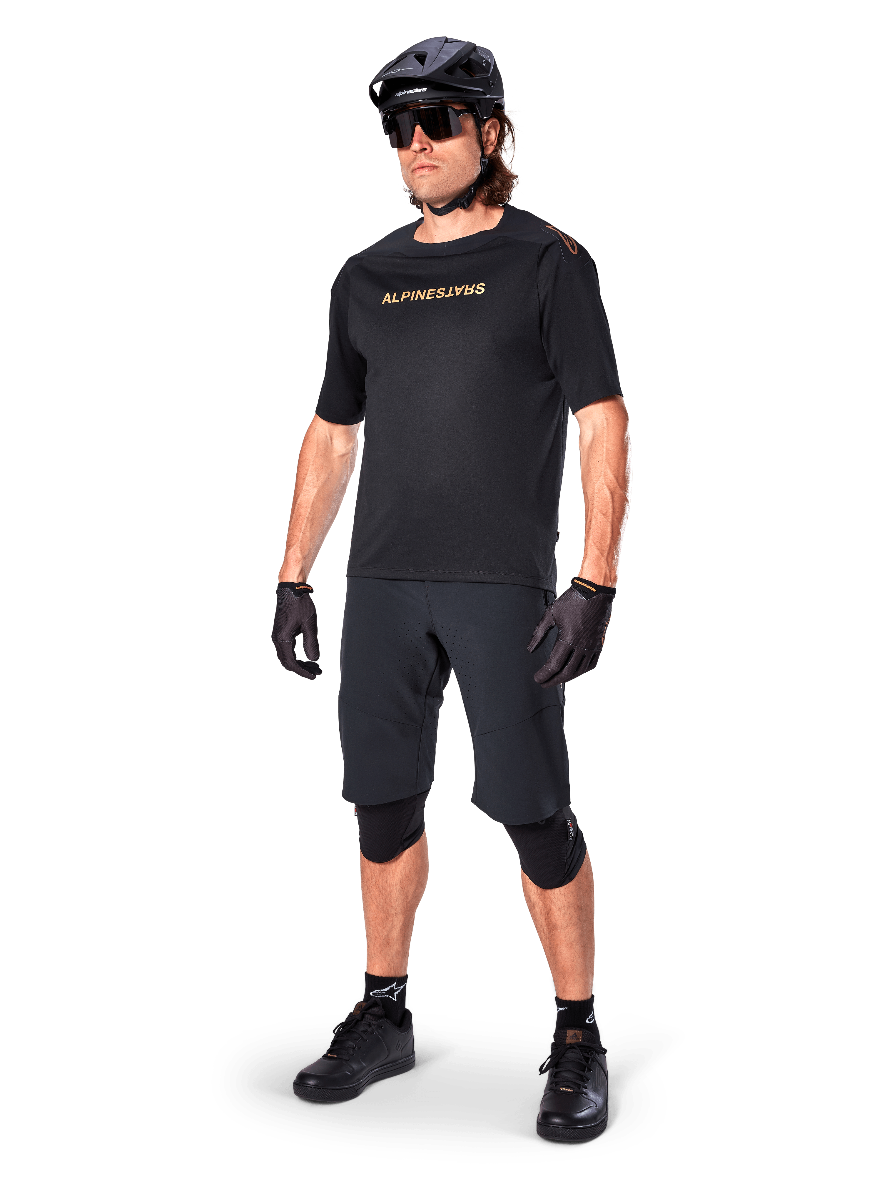 Cyklist iført Alpinestars A-Aria Elite shorts, sorte mountainbike-shorts, kombineret med en matchende sort trøje med guld-branding, sort hjelm, solbriller og knæbeskyttere, komplet performance-outfit til terrænkørsel