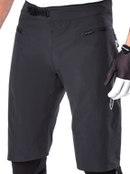 Alpinestars A-Aria Shorts, mountainbike-shorts, sort, nærbillede af taljen og lårene foran, der viser ratchet-lukkesystemet og lynlåslommer, bæres med sort-hvide cykelhandsker, performance trail-udstyr.
