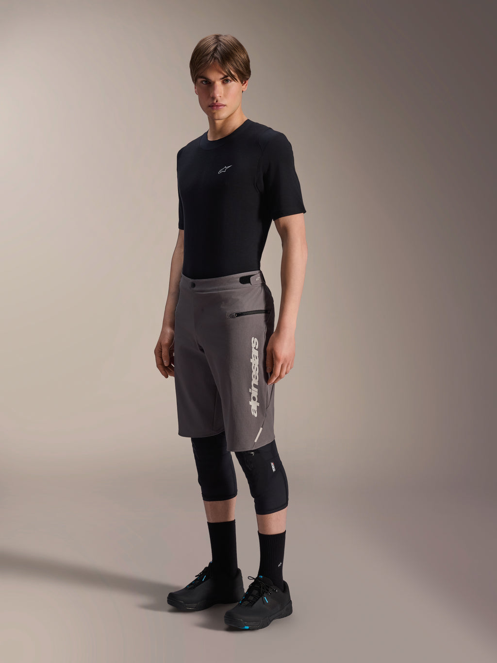 Alpinestars A-Dura Elite Shorts, Dark Gray mountain bike shorts, worn by a model with a black t-shirt and knee guards, featuring a vertical white Alpinestars logo on the left leg, technical Cordura fabric for off-road cycling.