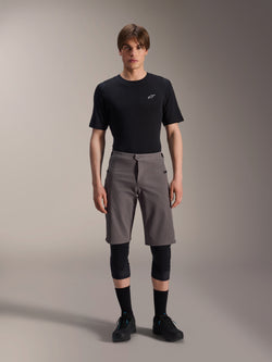 Alpinestars A-Dura Elite Shorts, Dark Gray Mountain Bike Shorts, worn with a black crew neck t-shirt and black knee guards, knee-length performance cycling apparel featuring a front snap closure and durable Cordura fabric.
