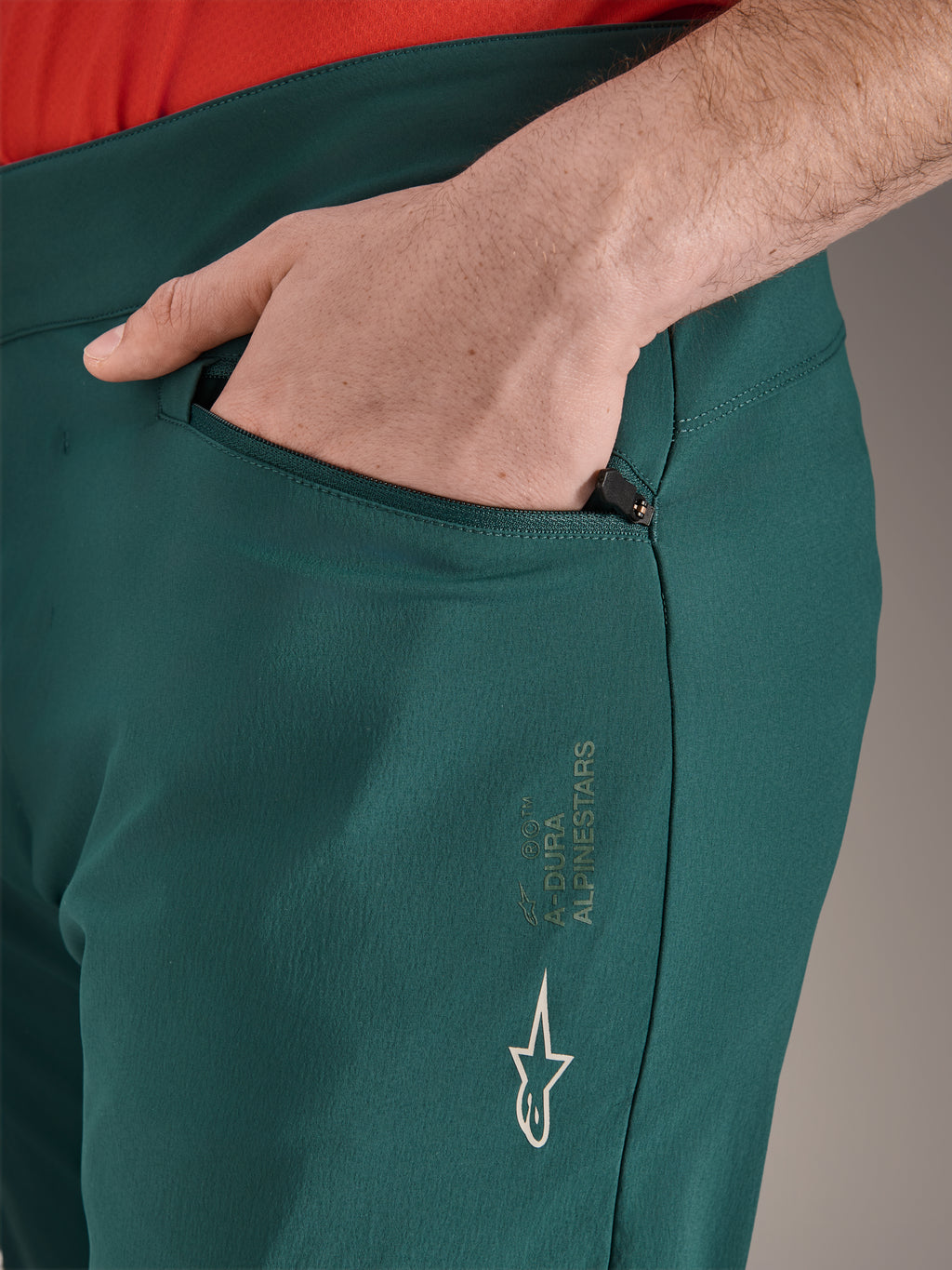 Alpinestars A-Dura bukser i Dark Pine grøn, tætbillede af lynlåslomme med en hånd, hvidt Alpinestars-logo og tekst på låret, slidstærke MTB-cykelbukser