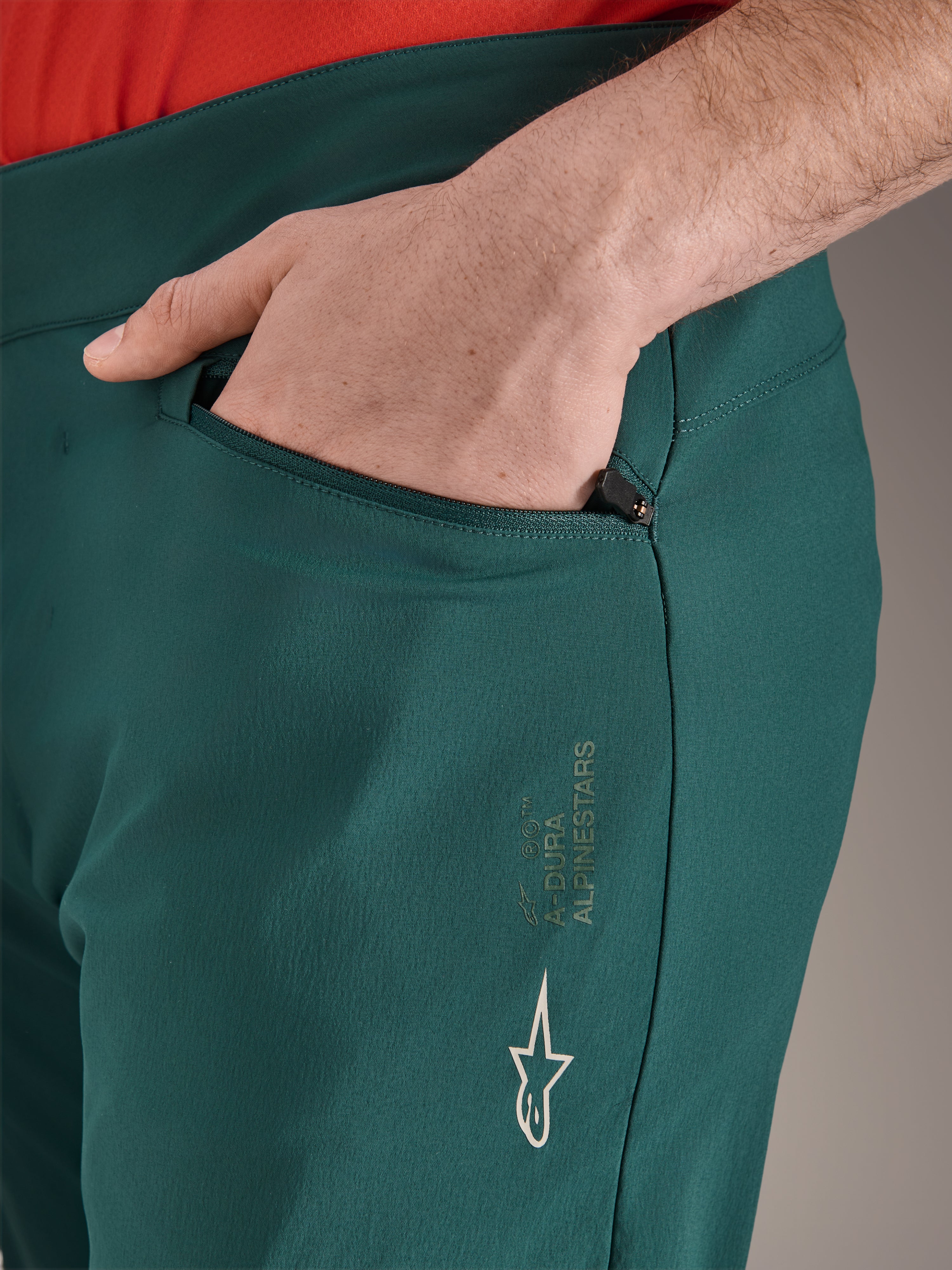 Alpinestars A-Dura bukser i Dark Pine grøn, tætbillede af lynlåslomme med en hånd, hvidt Alpinestars-logo og tekst på låret, slidstærke MTB-cykelbukser