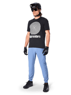 Alpinestars A-Dura bukser, MTB-bukser, Infinity Blue, båret med sort grafisk t-shirt med hvidt spiral-logo, sort hjelm, solbriller og sorte handsker, professionelt mountainbike-udstyr