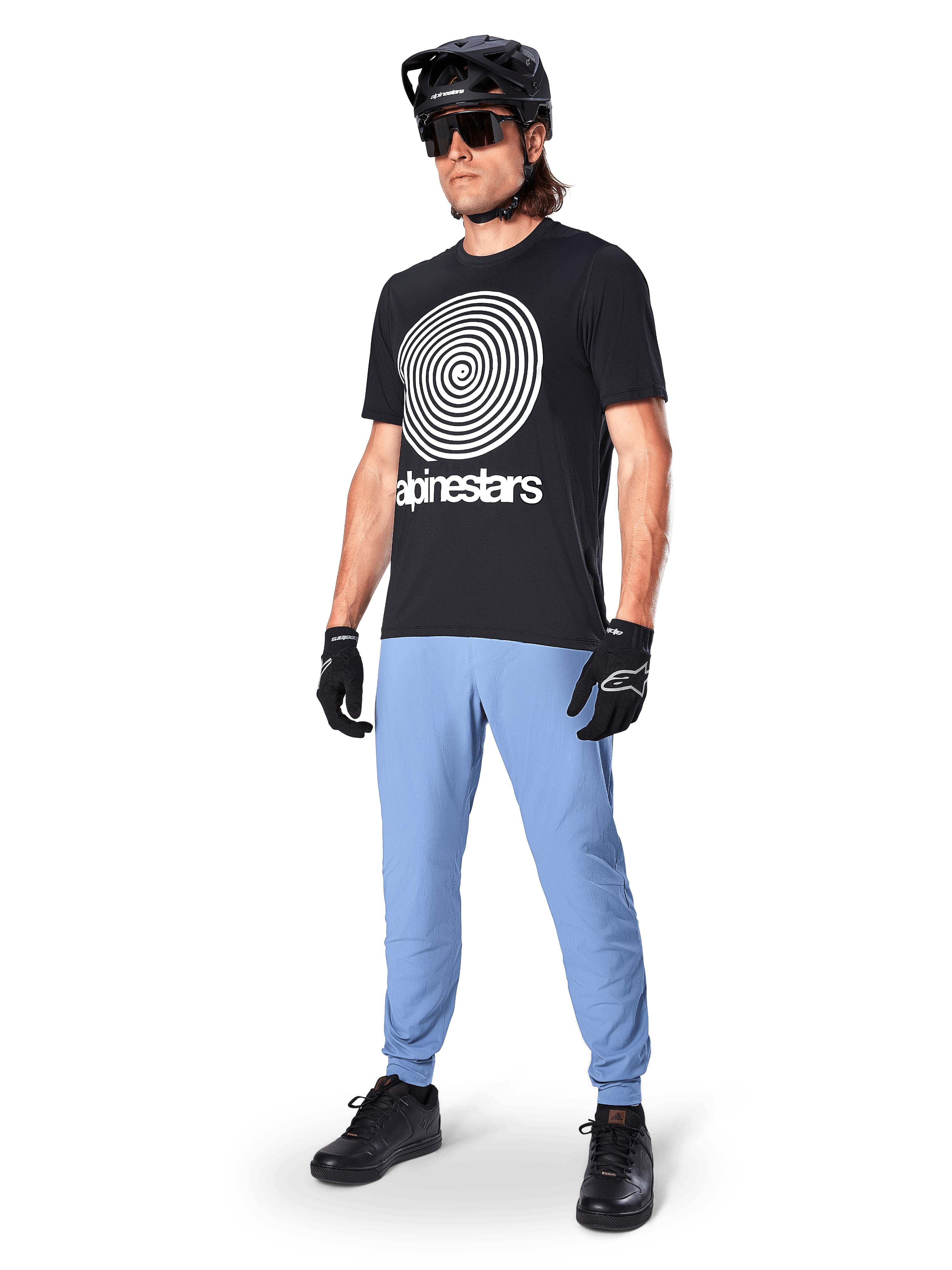 Alpinestars A-Dura bukser, MTB-bukser, Infinity Blue, båret med sort grafisk t-shirt med hvidt spiral-logo, sort hjelm, solbriller og sorte handsker, professionelt mountainbike-udstyr