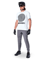 Alpinestars A-Dura bukser, MTB-bukser, mørkegrå, båret med hvid t-shirt med spiralgrafik, sort MTB-hjelm, solbriller og sorte handsker, holdbart 2-vejs strækstof til terrænkørsel