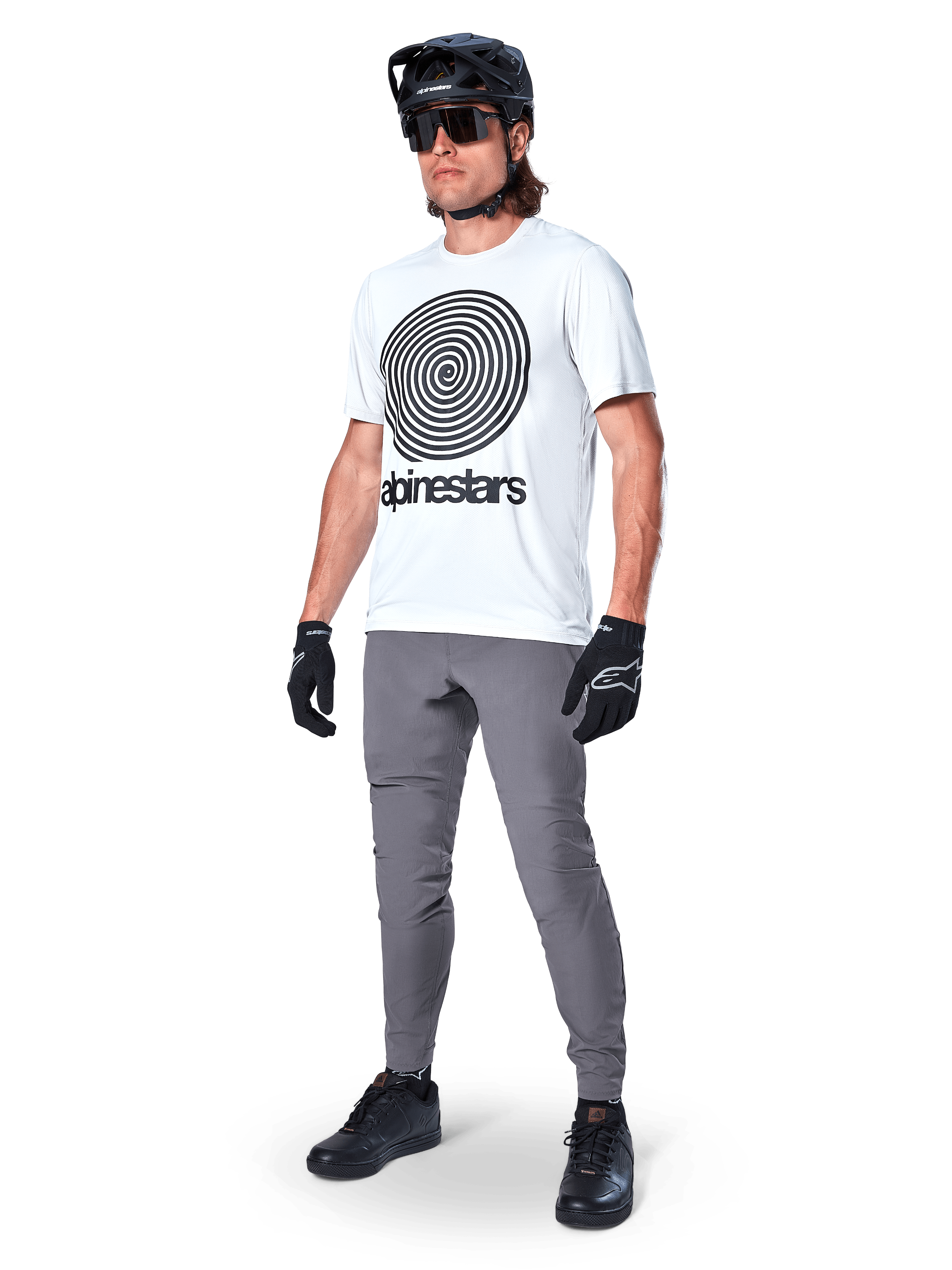 Alpinestars A-Dura bukser, MTB-bukser, mørkegrå, båret med hvid t-shirt med spiralgrafik, sort MTB-hjelm, solbriller og sorte handsker, holdbart 2-vejs strækstof til terrænkørsel