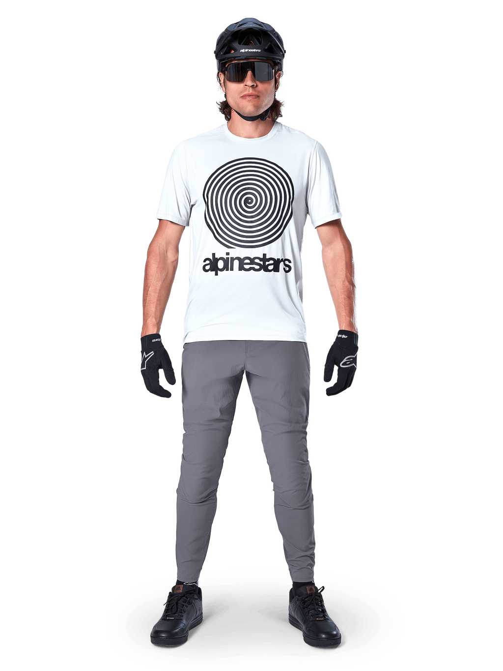 Alpinestars A-Dura bukser, MTB-cykelbukser, mørkegrå, båret med en hvid grafisk t-shirt, sort hjelm og handsker, med et holdbart 2-vejs strækdesign og almindelig pasform til mountainbiking