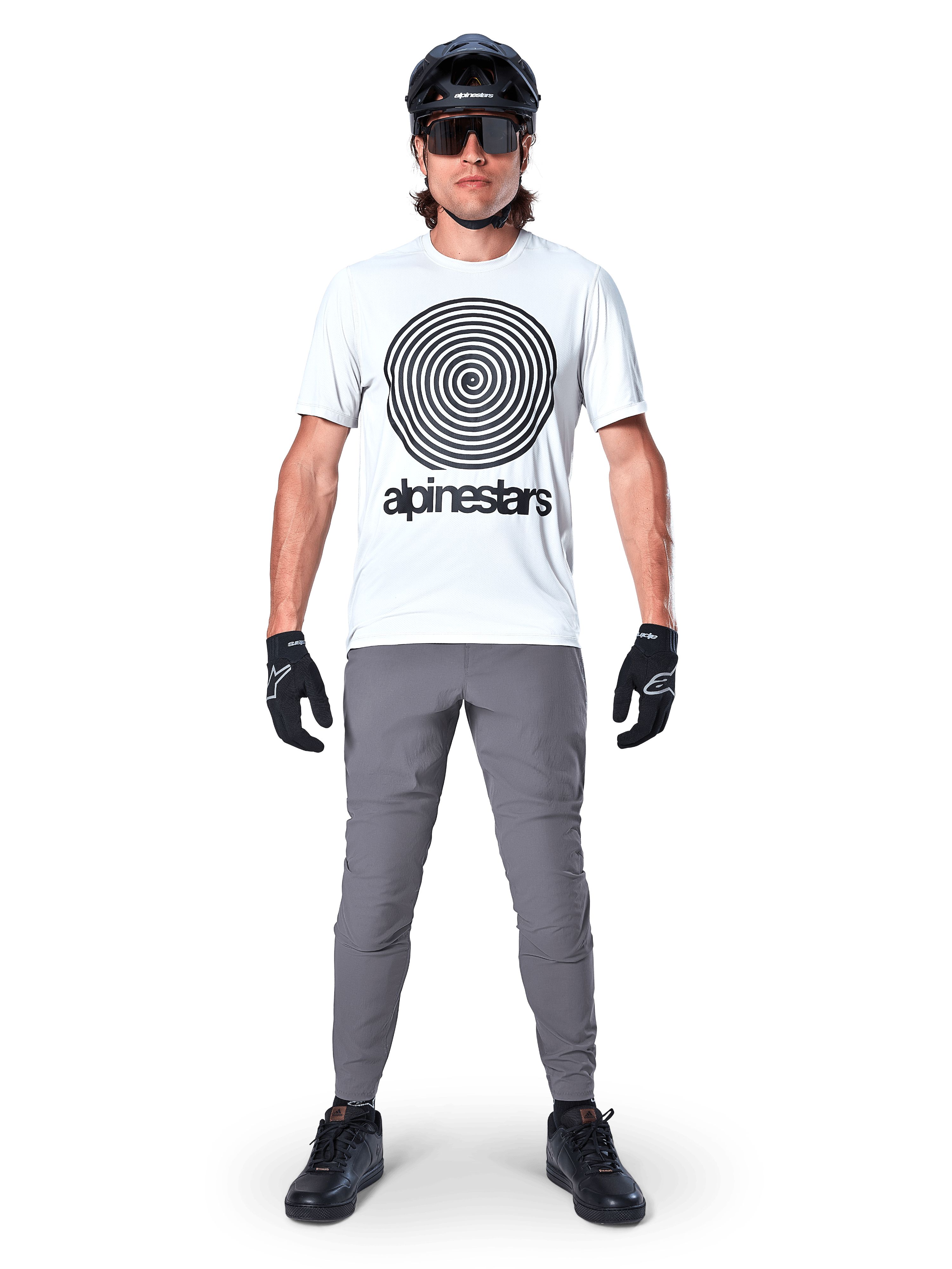 Alpinestars A-Dura bukser, MTB-cykelbukser, mørkegrå, båret med en hvid grafisk t-shirt, sort hjelm og handsker, med et holdbart 2-vejs strækdesign og almindelig pasform til mountainbiking