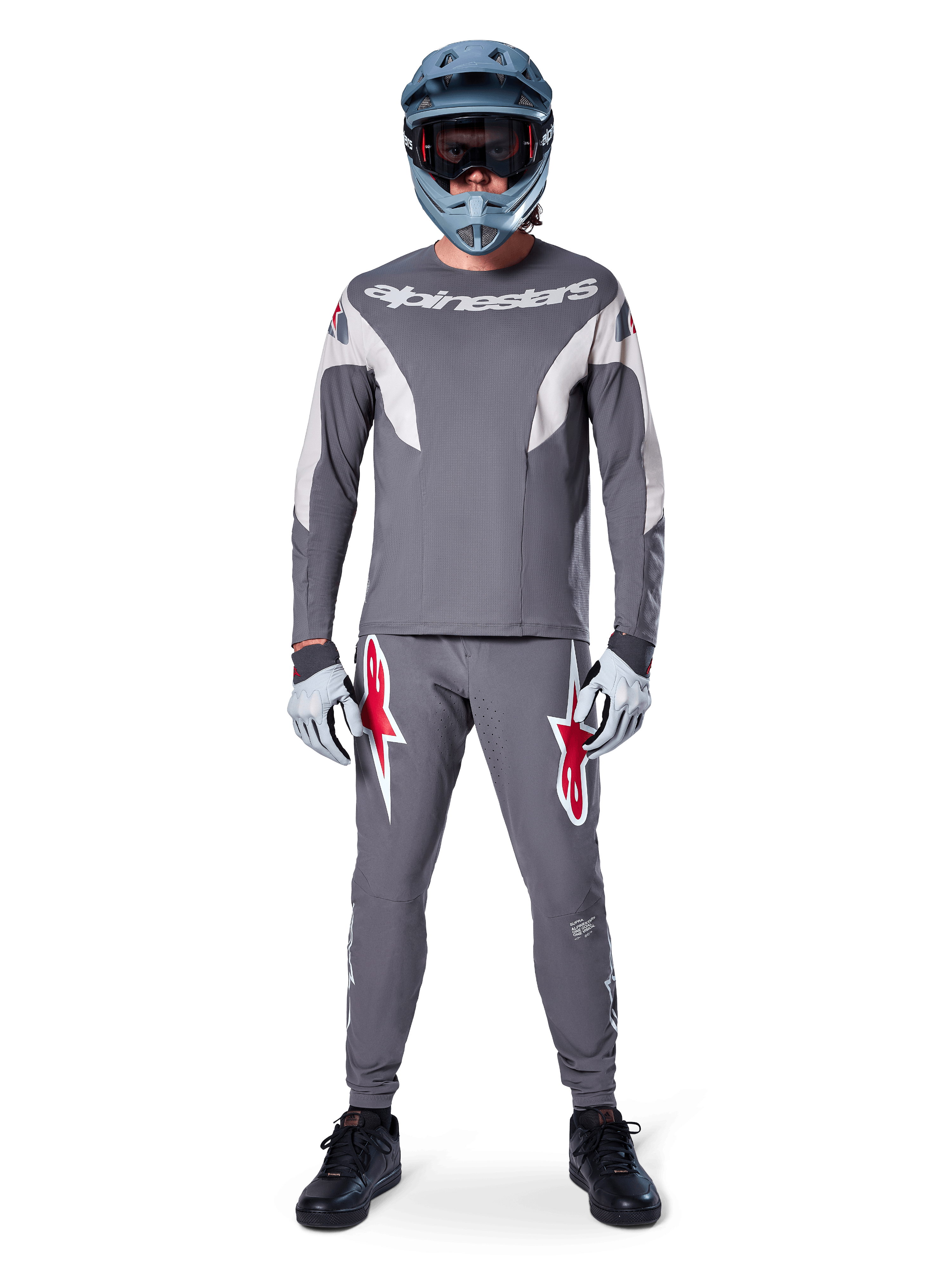 MTB-rytter iført Alpinestars A-Supra Race bukser i mørkegrå, med markante røde logo-detaljer på lårene, matchende grå jersey, blå full-face hjelm og sorte sko, komplet off-road racersæt