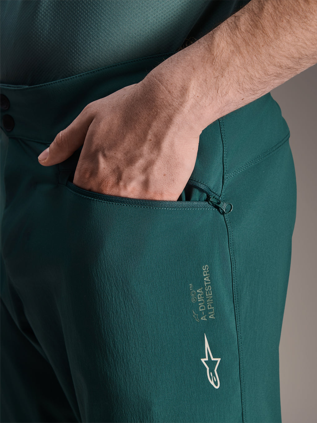 Alpinestars A-Dura shorts i Dark Pine, nærbillede af hånd i mesh-foret lynlåslomme, hvidt Alpinestars stjerne-logo og A-Dura branding, slidstærk tekstilkonstruktion til mountainbiking