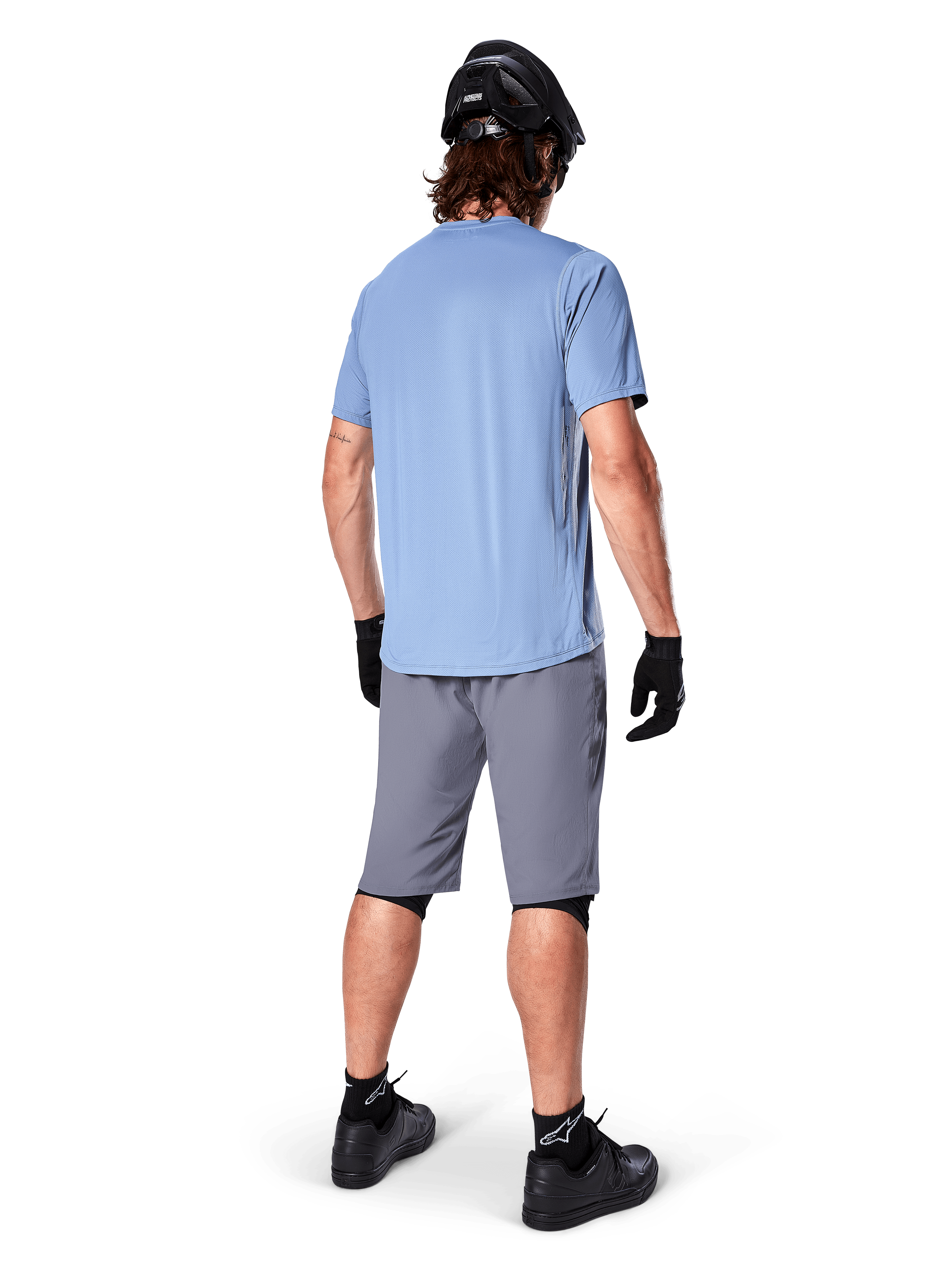 Alpinestars A-Dura shorts, mørkegrå MTB-shorts, set bagfra, båret med en lyseblå kortærmet trøje og sort mountainbike-hjelm, sorte handsker og strømper med sorte sko, funktionelt og holdbart gear til terrænkørsel